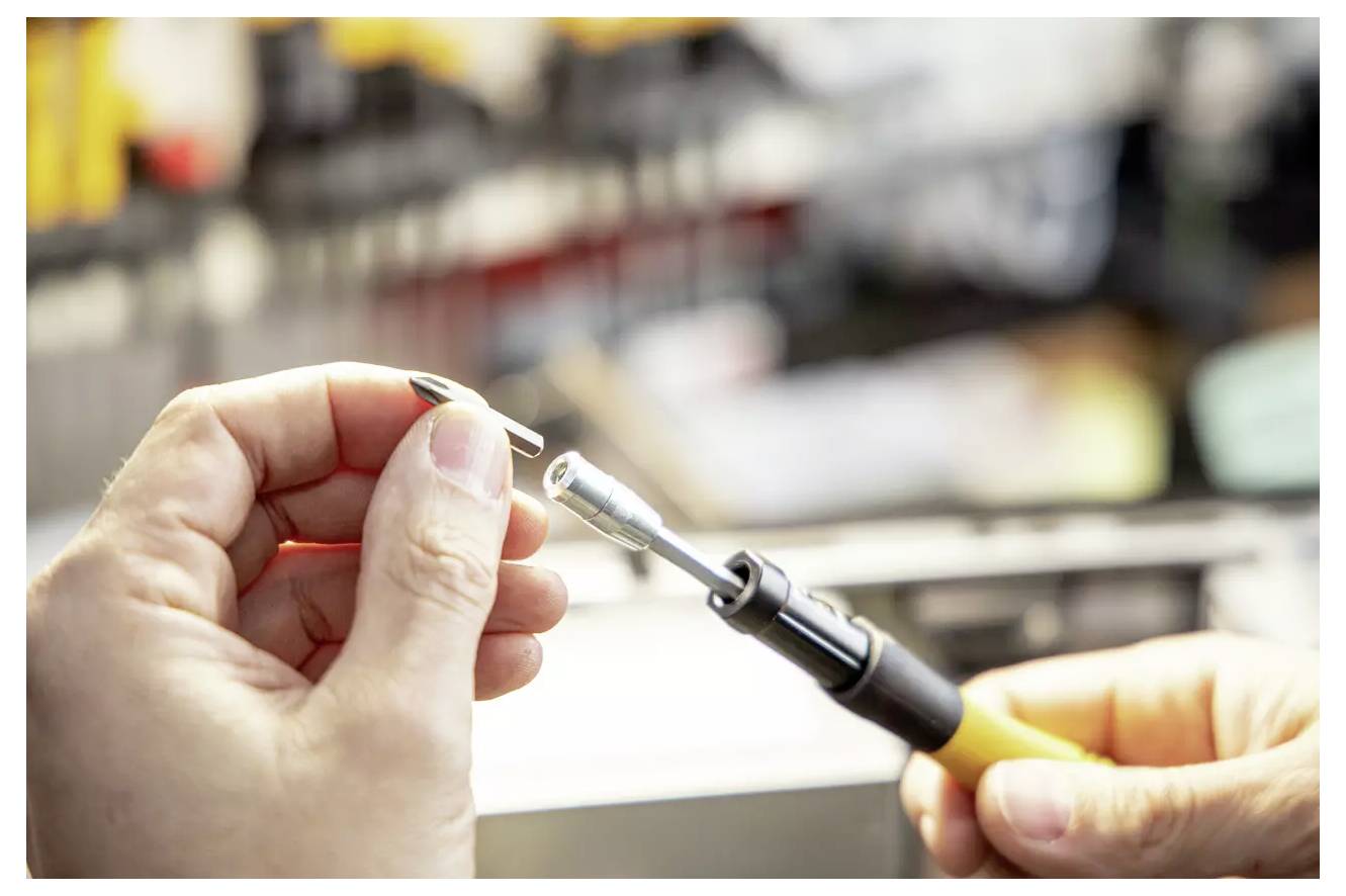 Eine Person hält ein Lötgerät mit einem kleinen Metallteil, was auf eine laufende Aufgabe der Präzisionsfertigung oder Elektronikreparatur hindeutet.