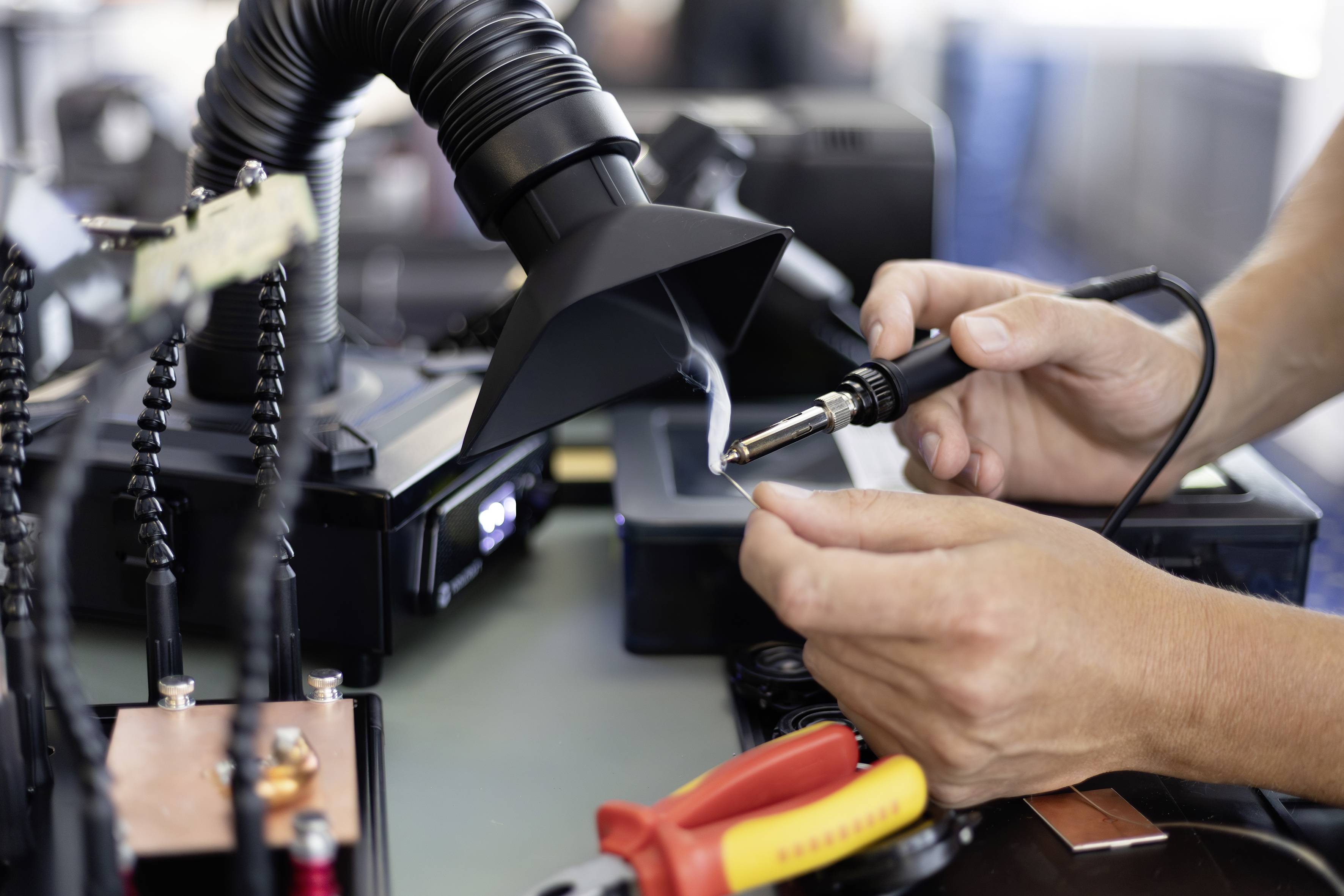 Hände, die einen Lötkolben verwenden, um eine Leiterplatte unter einem Absauggerät zu reparieren, mit Elektronikwerkzeugen auf der Werkbank.