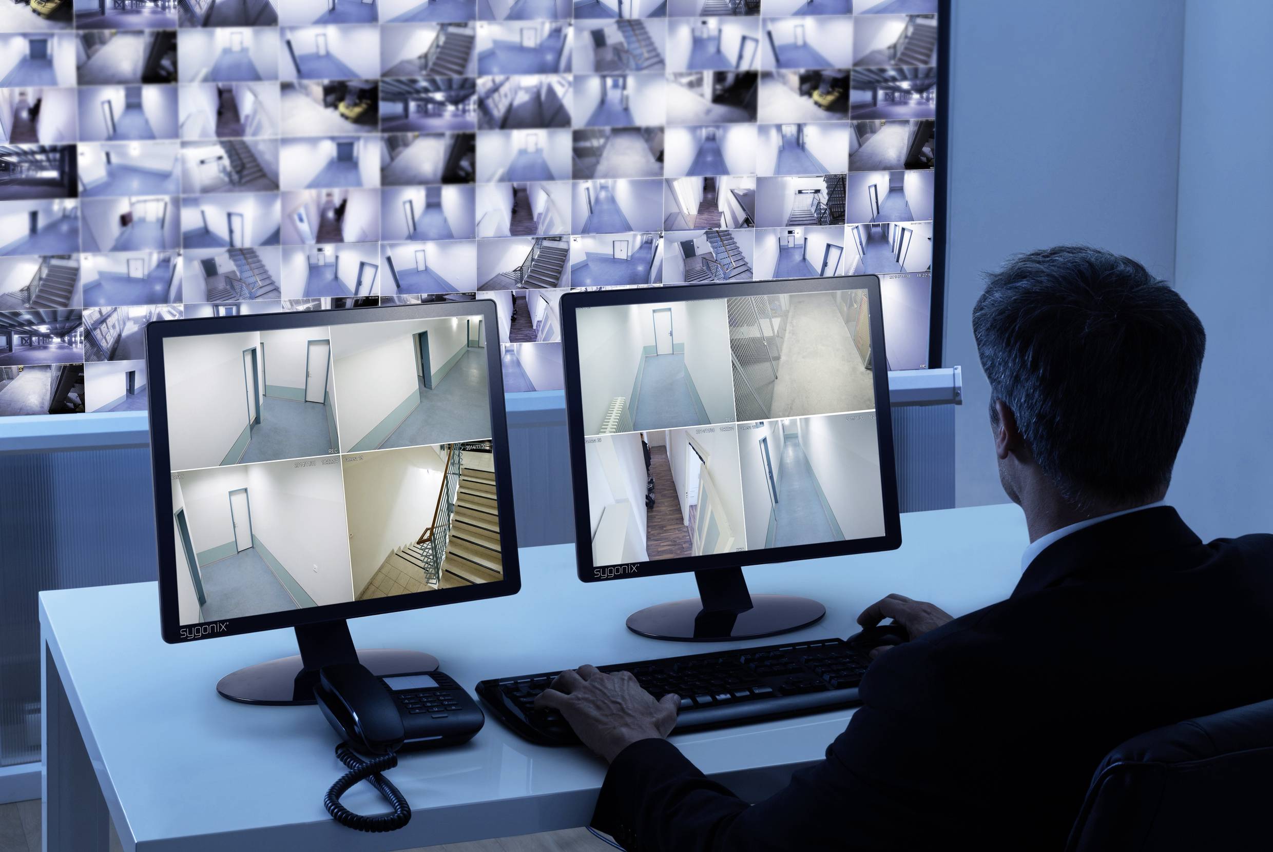 Ein Mann überwacht mehrere Überwachungskameras auf zwei Monitoren, die verschiedene Gebäudebereiche zeigen. Im Hintergrund viele Kamerabilder.
