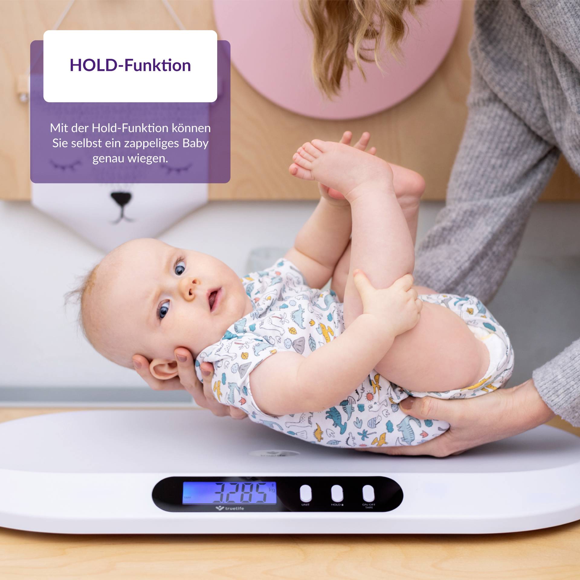 Eine Person wiegt ein Baby auf einer digitalen Waage. Eine Anzeige zeigt 7.85 kg. Ein Schild erklärt die 'HOLD-Funktion'.
