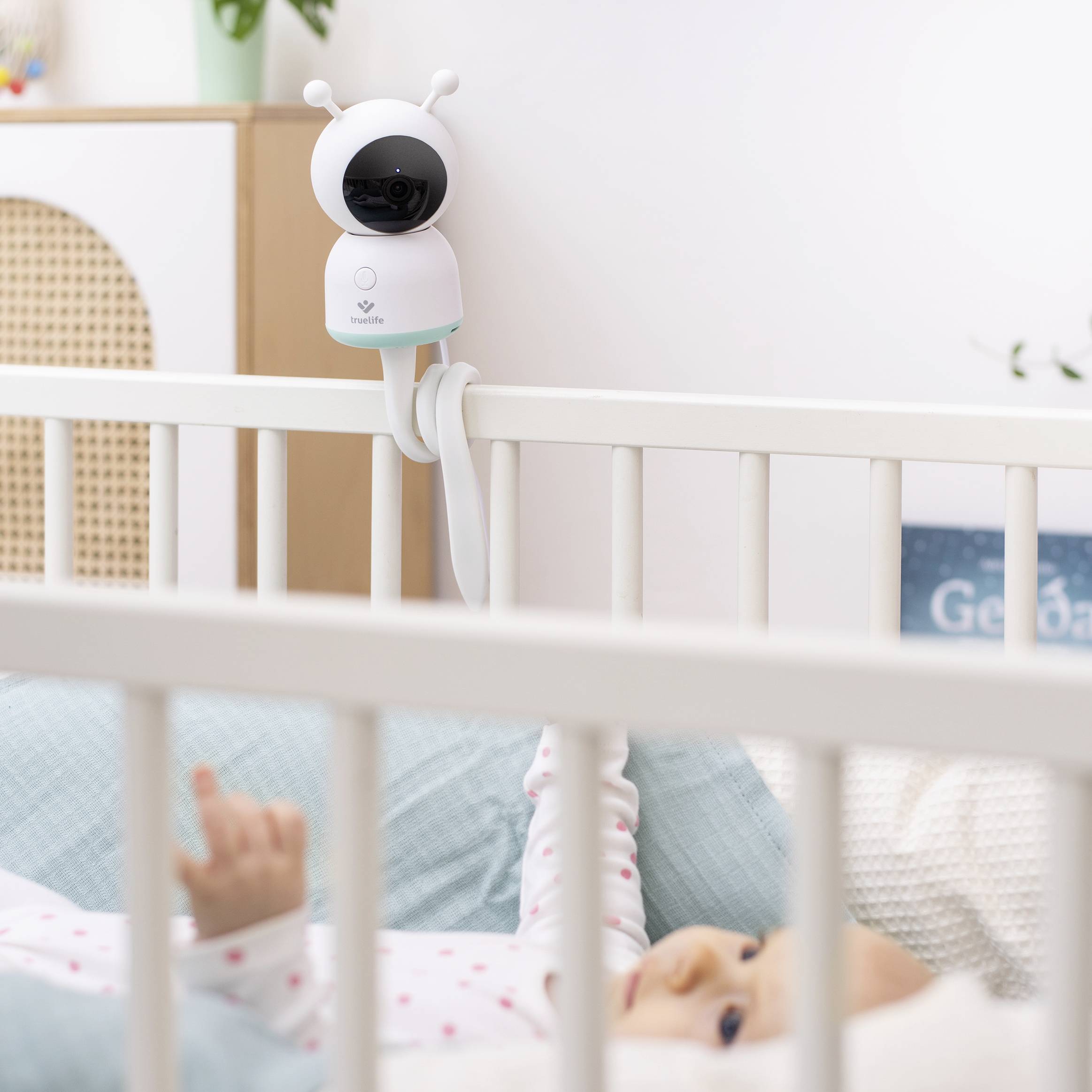 Eine Babyüberwachungskamera ist am Gitter eines Kinderbettes befestigt. Ein Baby liegt darunter und schaut nach oben.