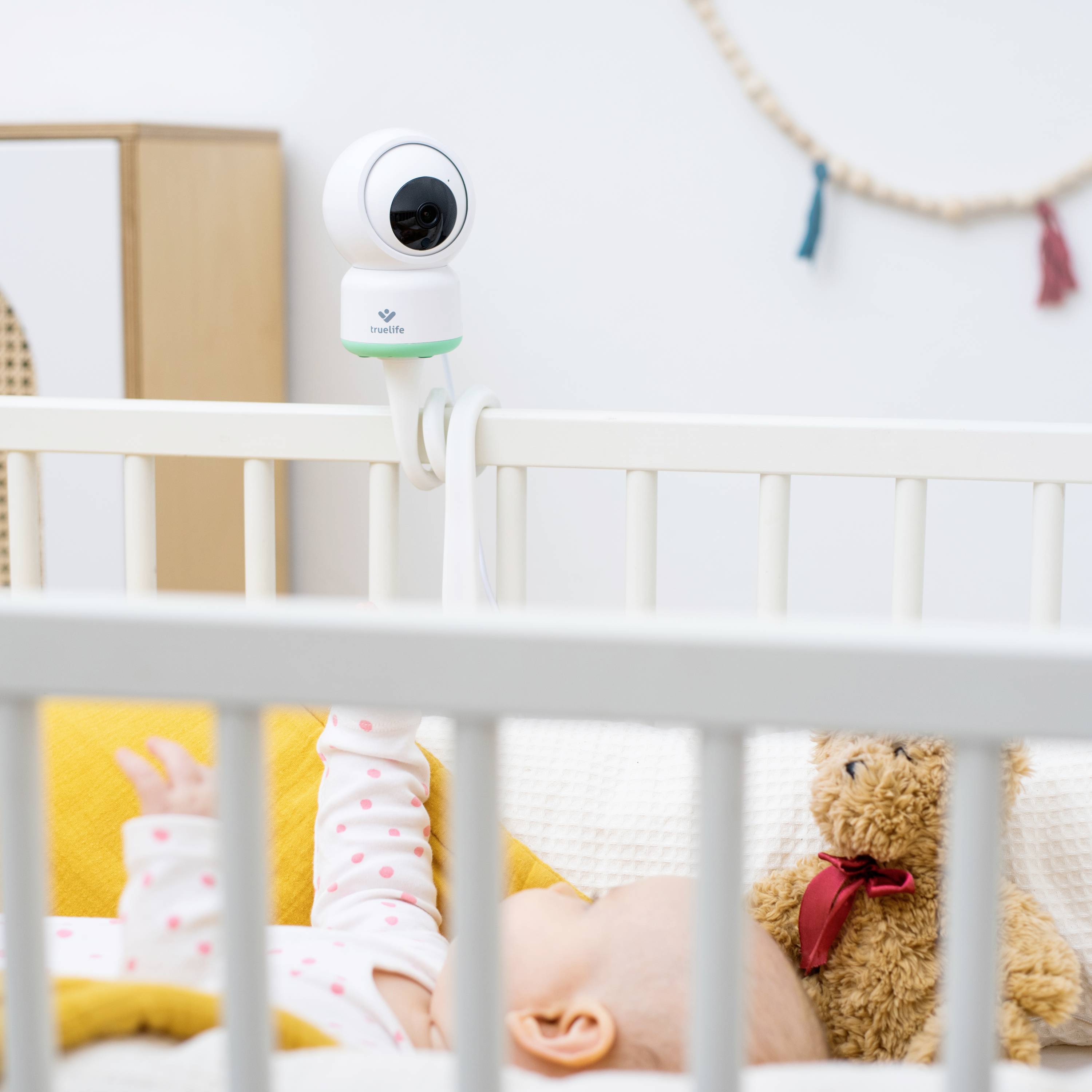 Ein Baby schläft in einem Kinderbett, während eine Kamera am Bett befestigt ist. Ein Plüschteddy liegt neben dem Baby.