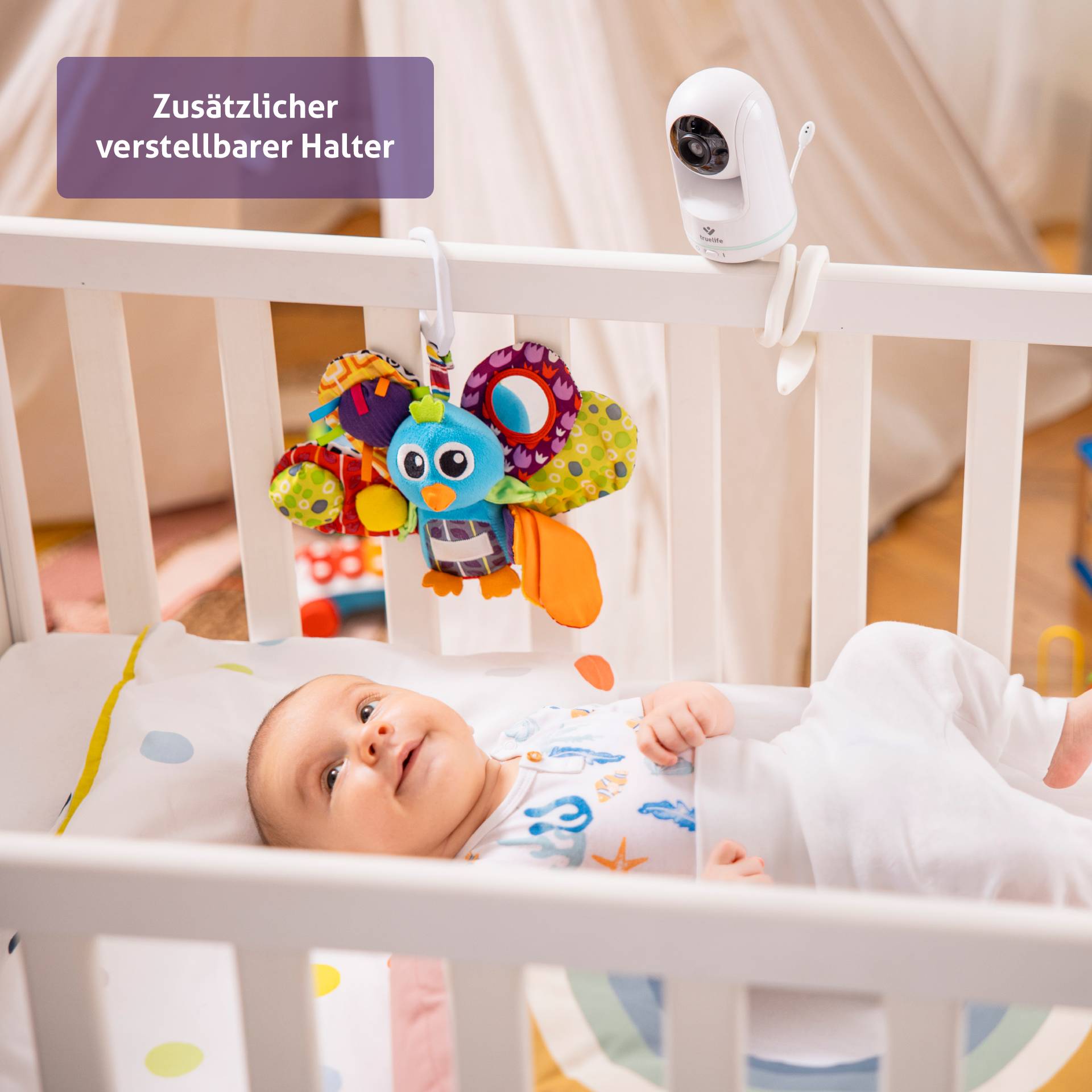 Ein Baby liegt in einem Bett, schaut lächelnd auf ein buntes Stoffspielzeug. Eine Kamera ist am Bett befestigt für Überwachung.