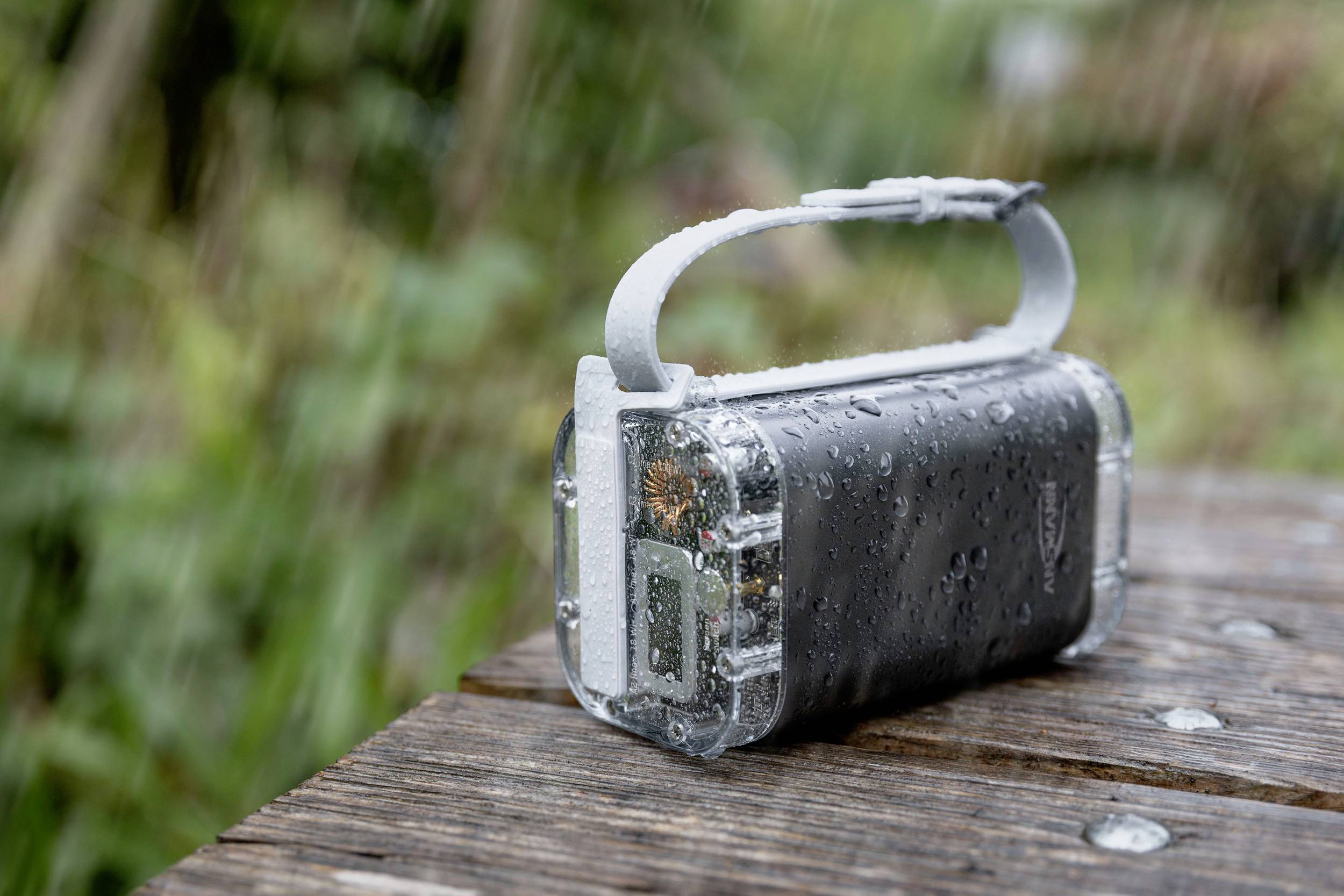 Ein wasserdichtes, tragbares Gerät mit Griff steht im Regen auf einem Holztisch. Es ist von Wassertropfen bedeckt.