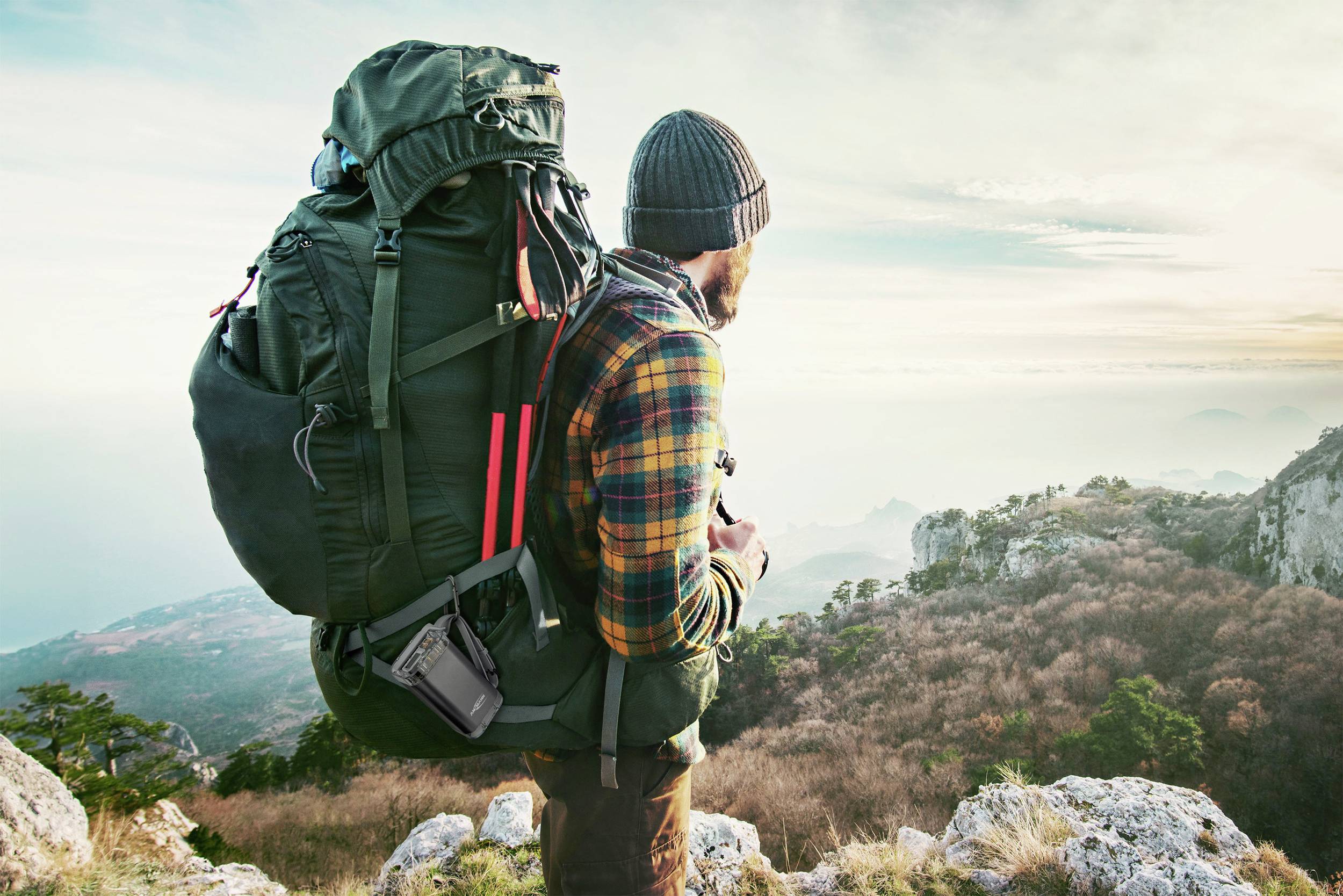 Eine Person mit großem Rucksack steht auf einem felsigen Berggipfel und blickt in die Ferne, während die Sonne scheint.