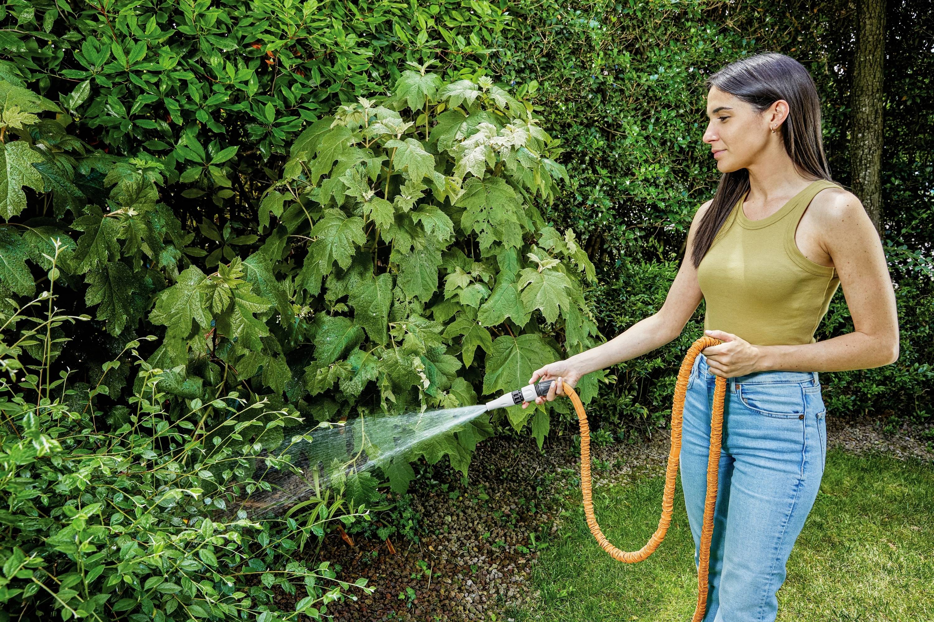 Frau gießt Pflanzen im Garten mit einem orangefarbenen Schlauch, umgeben von dichtem grünen Laub und Rasen.