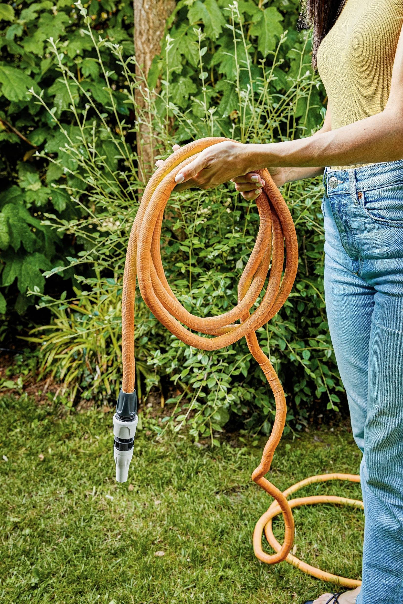 Eine Person hält einen orangefarbenen Gartenschlauch im Garten. Im Hintergrund sind grüne Büsche zu sehen.
