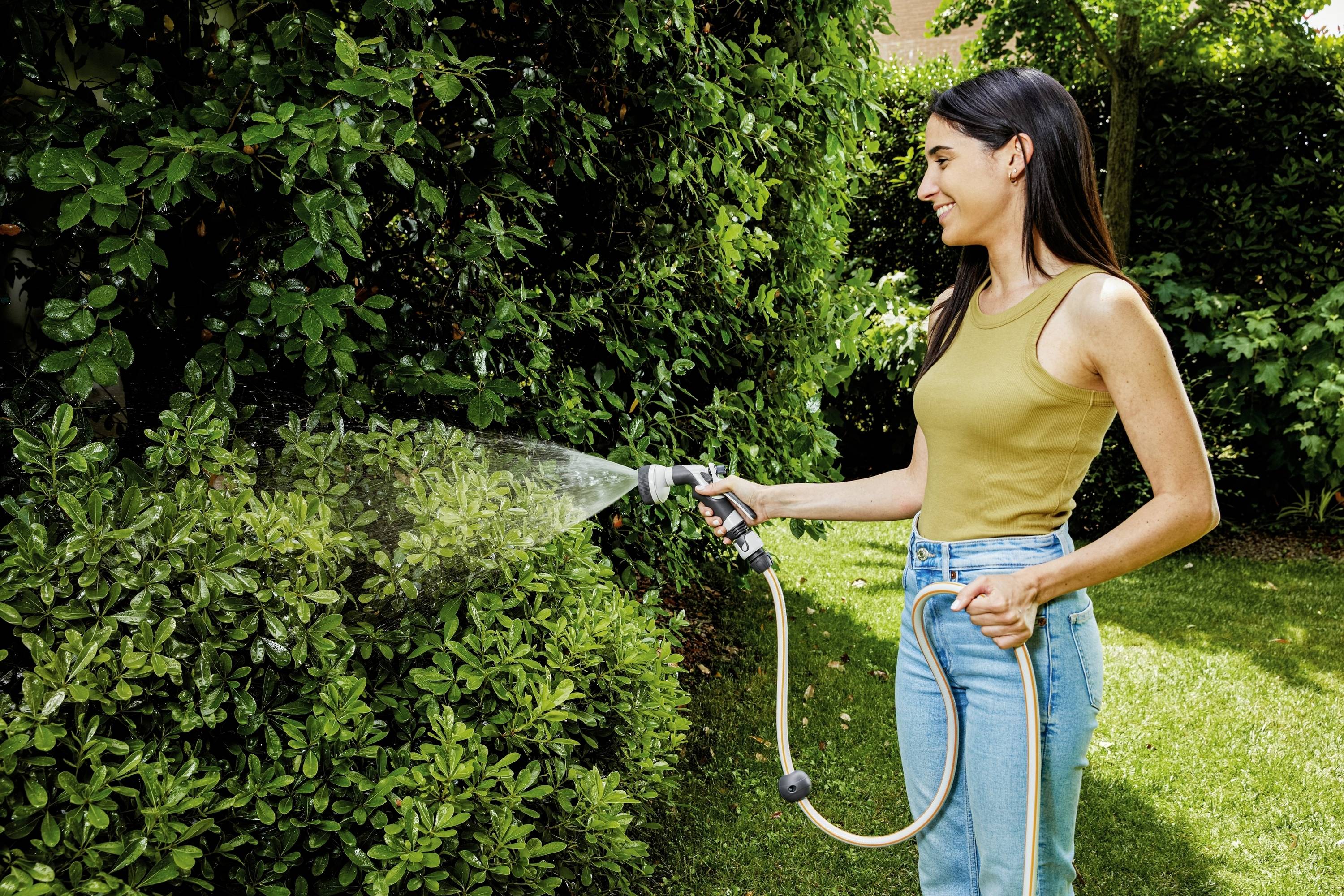 Eine Frau steht im Garten und gießt mit einem Wasserschlauch Büsche. Sie trägt ein gelbes Top und blaue Jeans. Es ist sonnig.