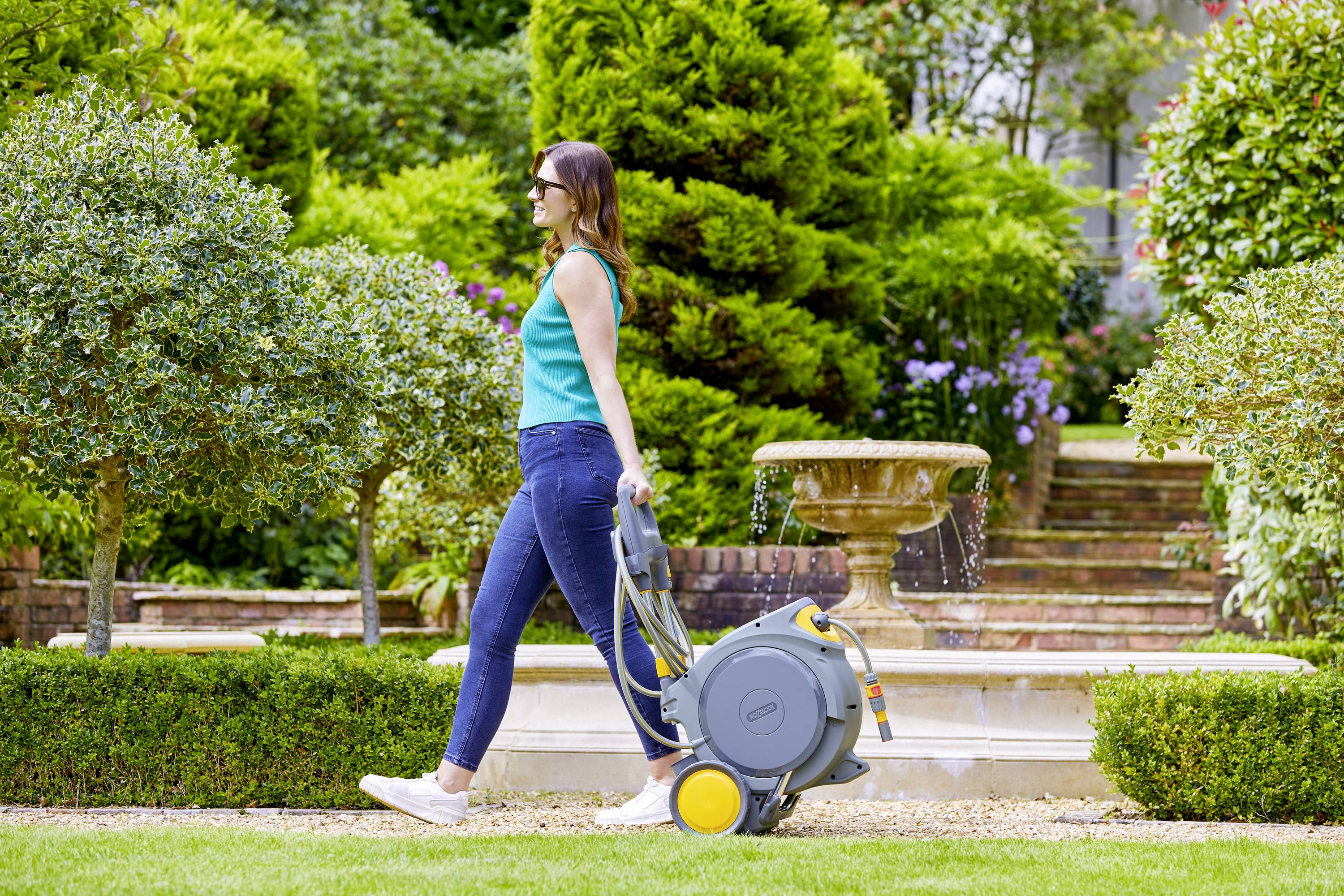 Eine Frau zieht einen Gartenschlauchwagen durch einen gepflegten Garten mit einem Springbrunnen im Hintergrund.