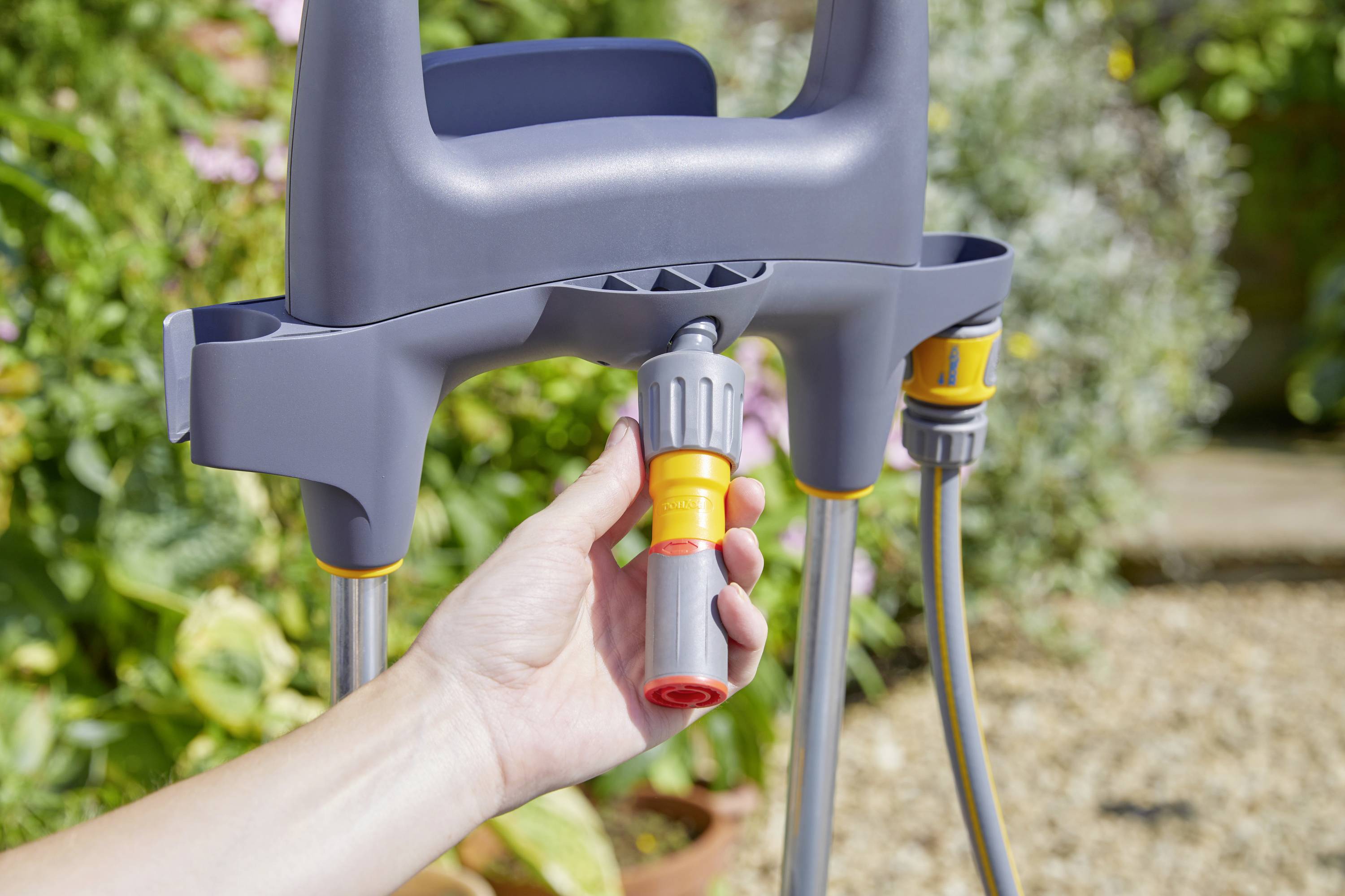 Eine Hand hält einen Schlauchverbinder, der mit einem Gartenschlauch und einer Halterung verbunden ist. Im Hintergrund sind Pflanzen zu sehen.