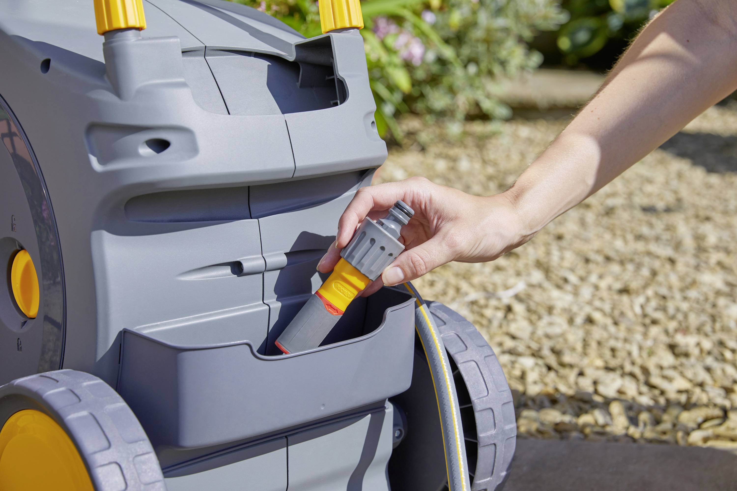 Hand steckt grauen Schlauchanschluss in die Halterung eines grauen Schlauchwagens. Kieselsteine und Pflanzen im Hintergrund.