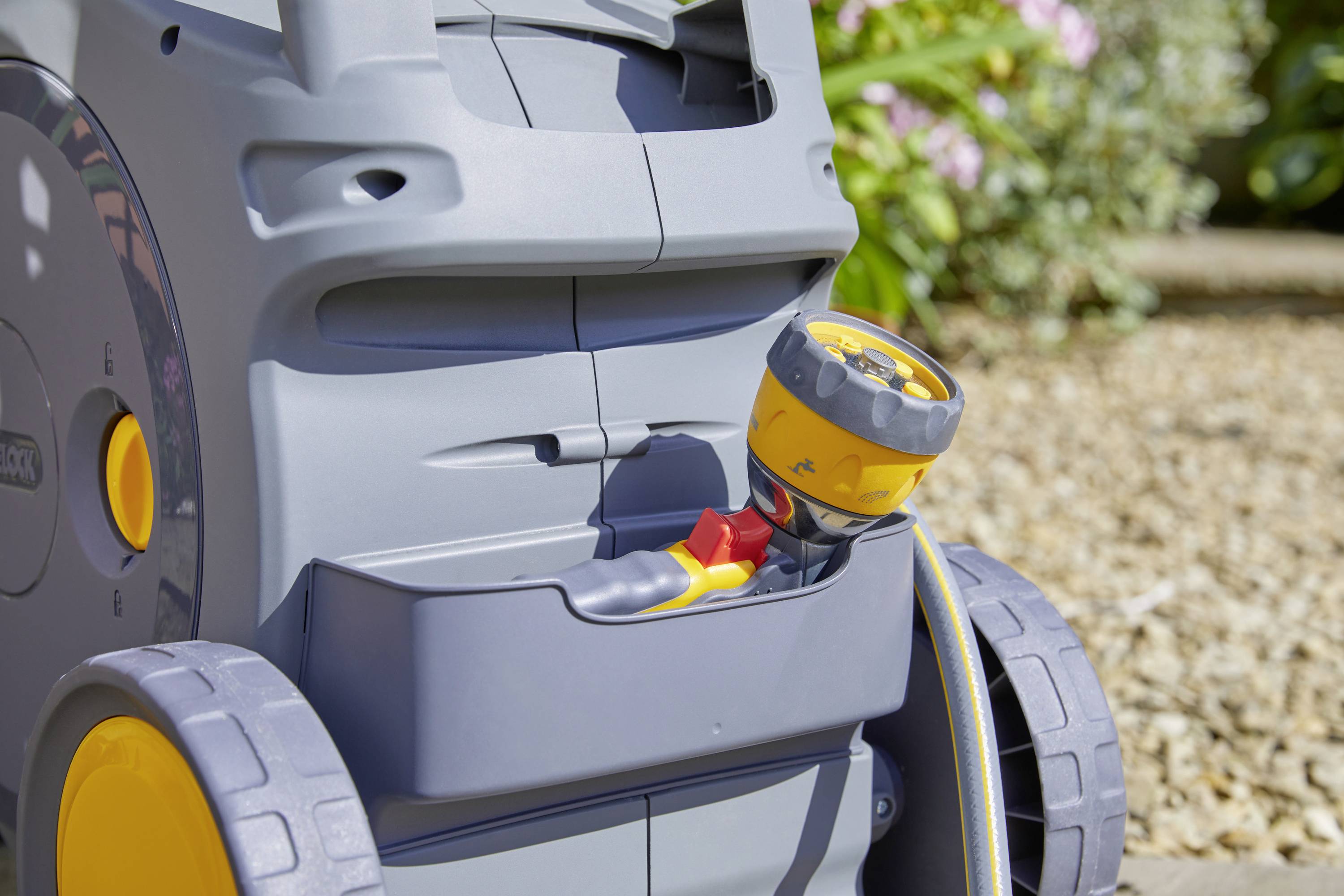 Gartenschlauchwagen mit gelber Düse und großen Rädern im Sonnenlicht auf Kiesboden.