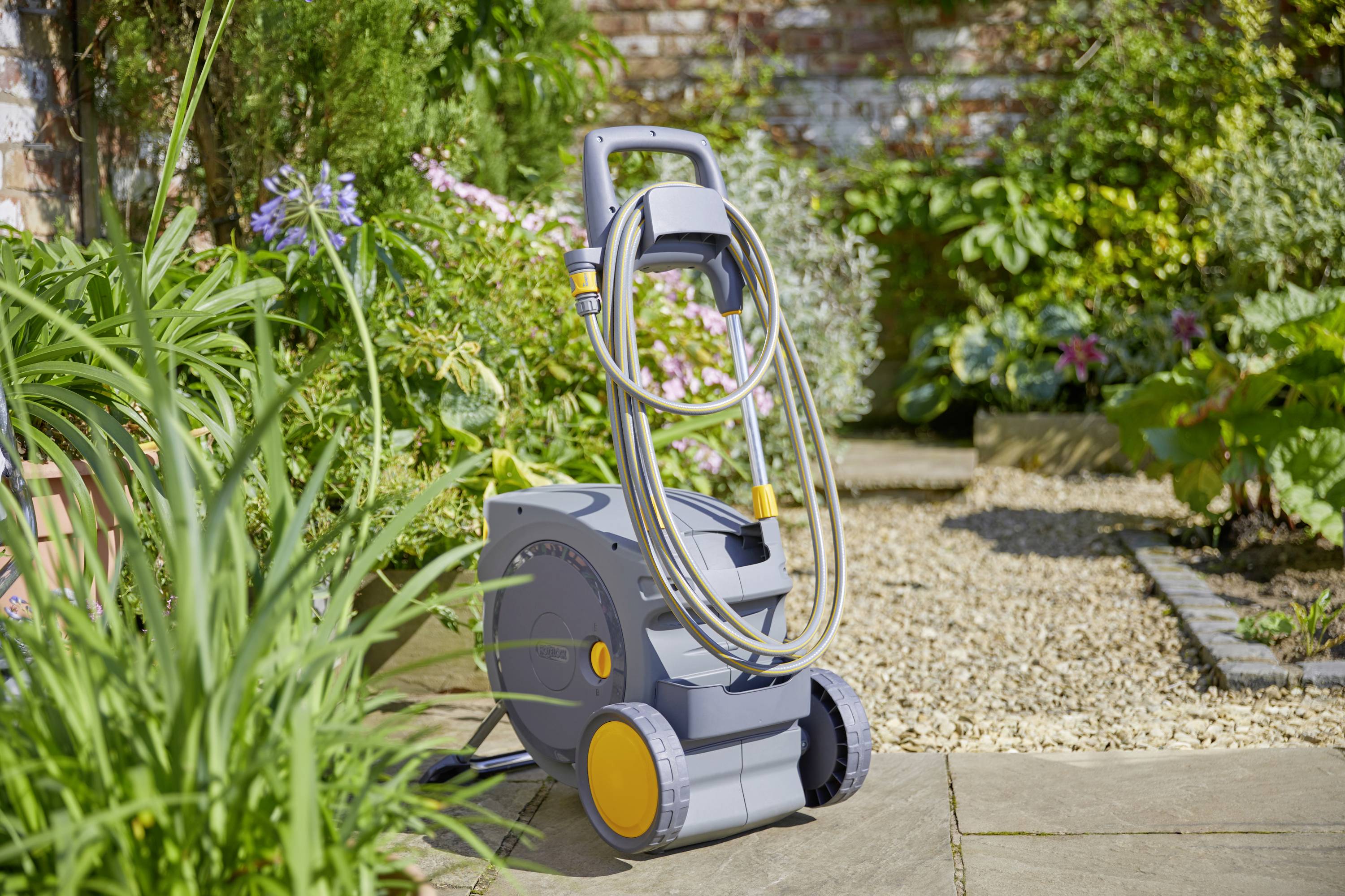 Ein mobiler Wasserschlauchwagen steht auf einer Terrasse im Garten, umgeben von grünen Pflanzen und Blumen. Ideal für die Gartenbewässerung.