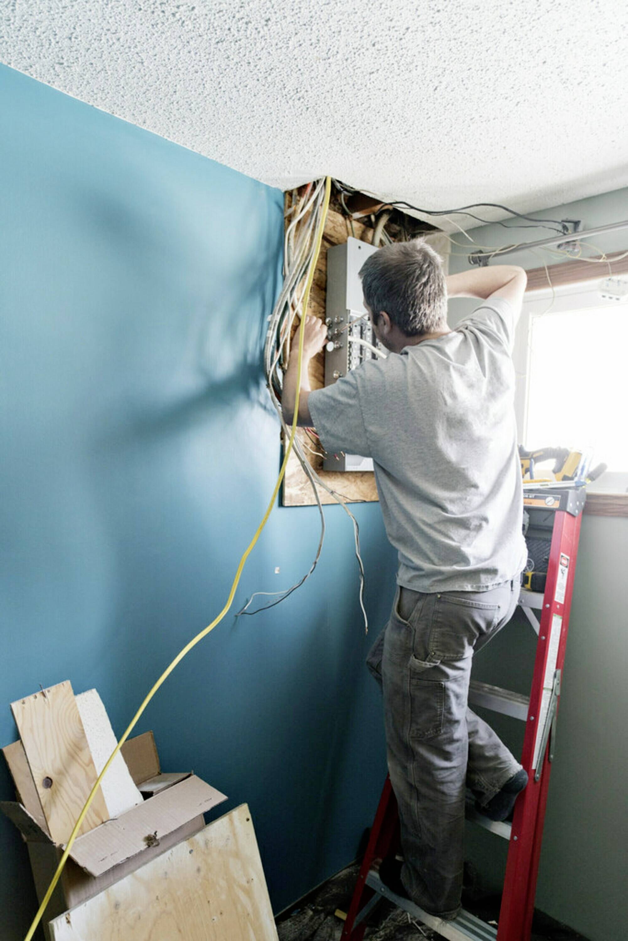 Eine Person repariert elektrische Kabel in einer Wand. Sie steht auf einer Leiter und arbeitet konzentriert, um den Schaden zu beheben.