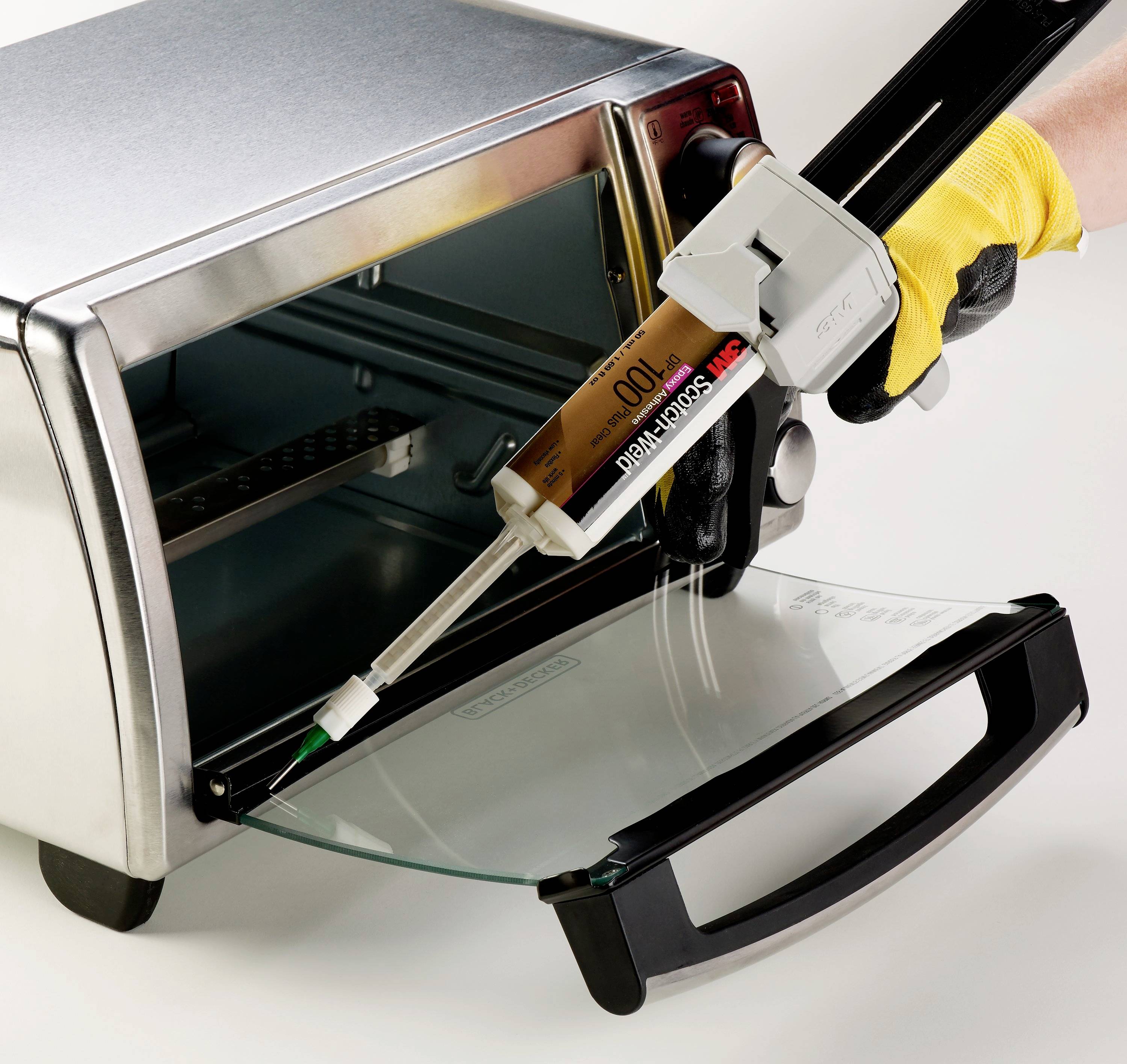 Eine Hand mit gelbem Handschuh hält eine Kartuschenpistole mit Metallkleber, der auf die offene Tür eines Toasters aufgetragen wird.