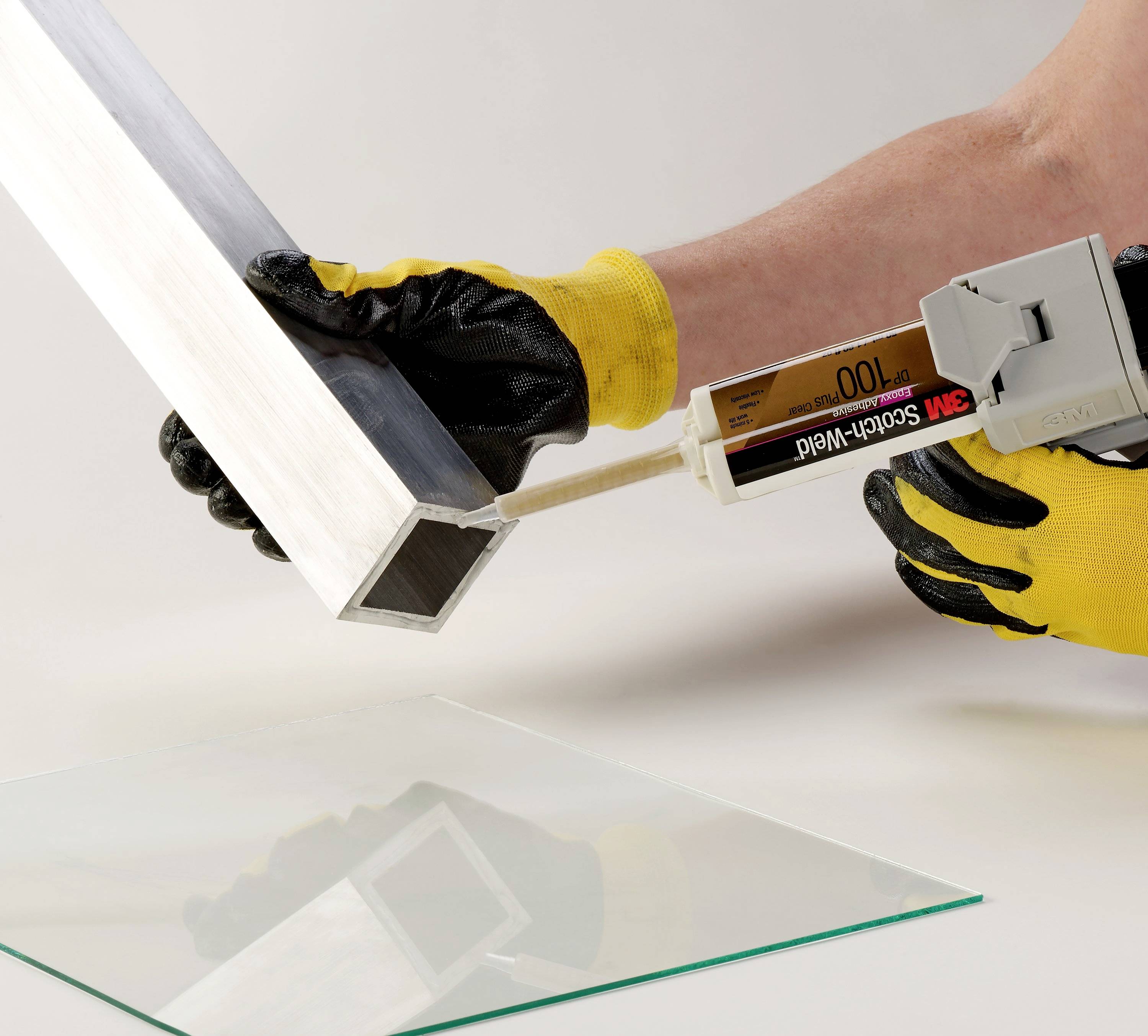 Eine Person mit gelben Handschuhen trägt mit einer Klebepistole Klebstoff auf die Kante eines Metallwinkels auf, daneben Glasplatte.