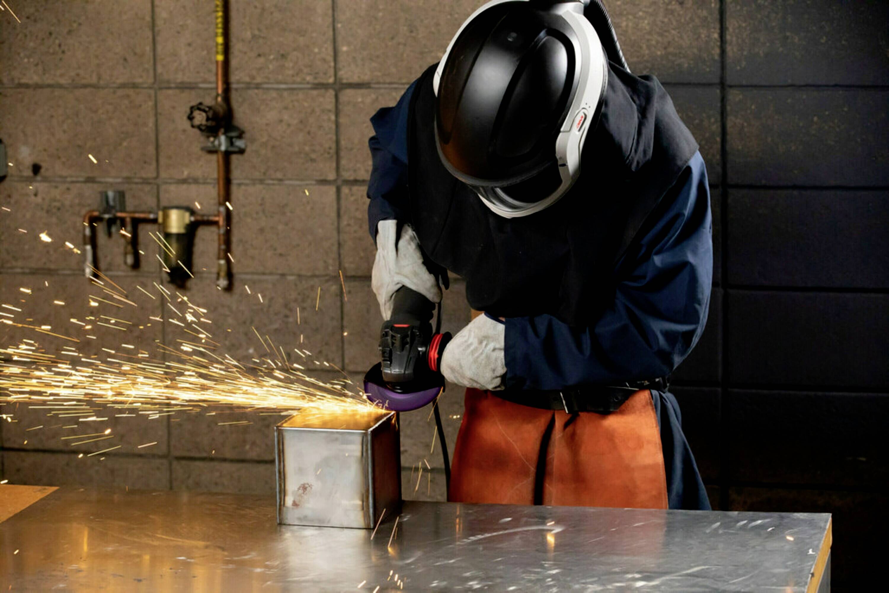 Eine Person schleift eine Metallbox mit einem Winkelschleifer. Funken fliegen, Schutzkleidung wird getragen. Schweißen im Arbeitskontext.