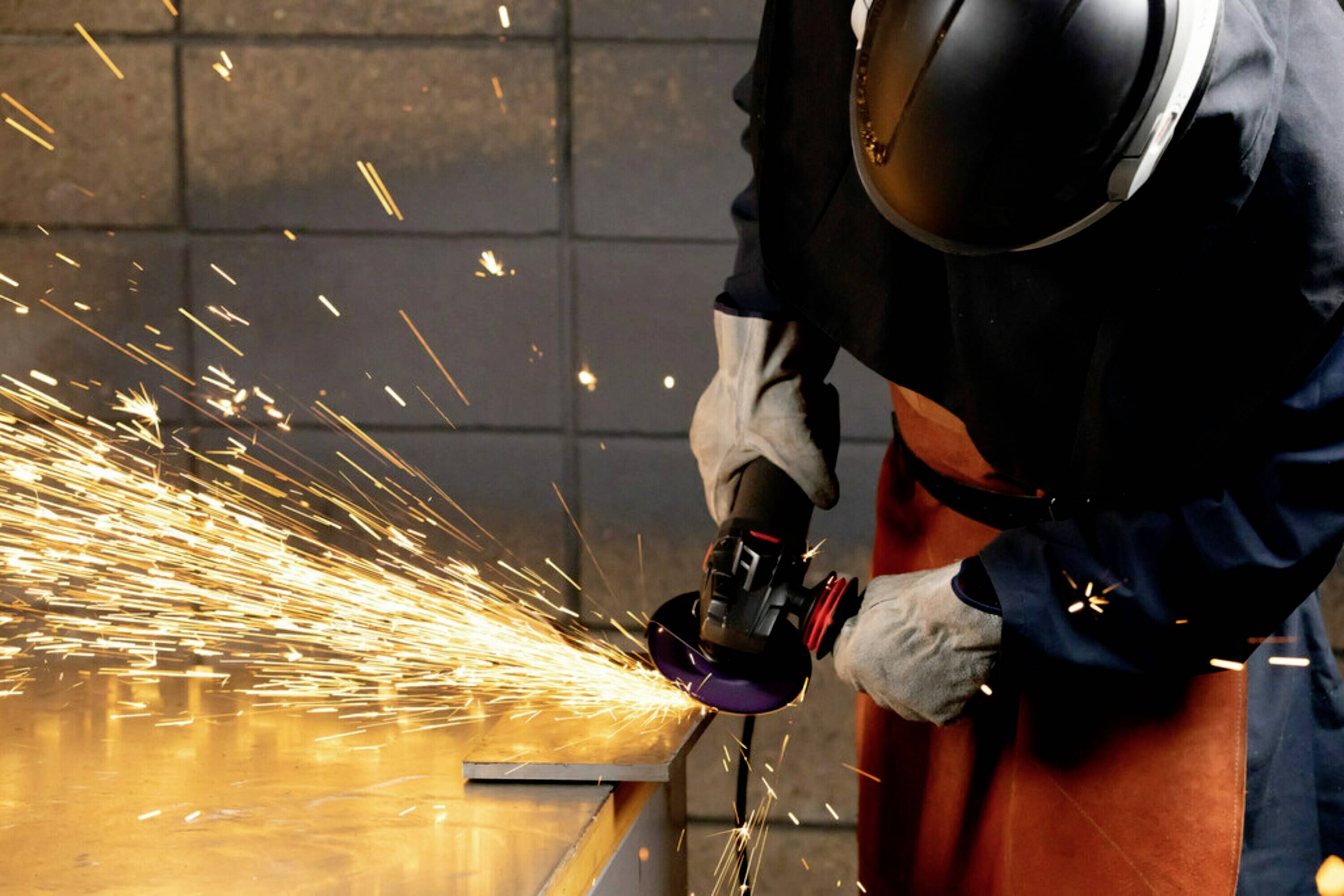 Eine Person schleift Metall mit einem Winkelschleifer, Funken sprühen. Sie trägt Schutzkleidung, einschließlich Helm und Handschuhe.