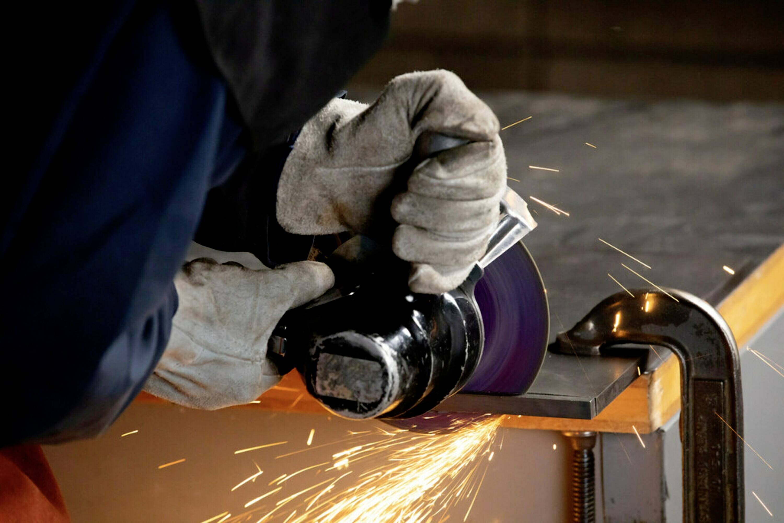Person schleift Metall mit einer Flex, Funken sprühen. Der Tisch ist mit einer Klemme gesichert. Trägt Handschuhe und Schutzkleidung.