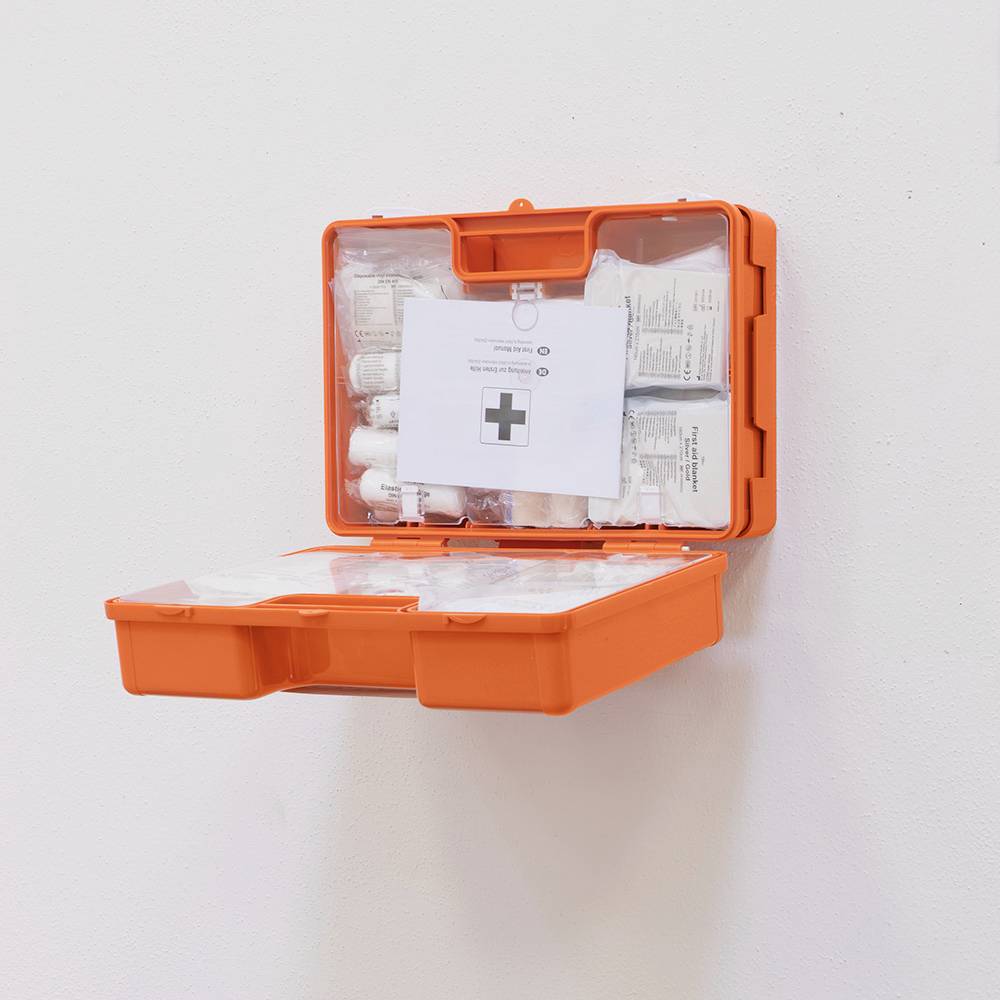 Ein geöffneter, orangefarbener Erste-Hilfe-Kasten an der Wand, gefüllt mit Verbänden, Pflastern und medizinischen Utensilien.