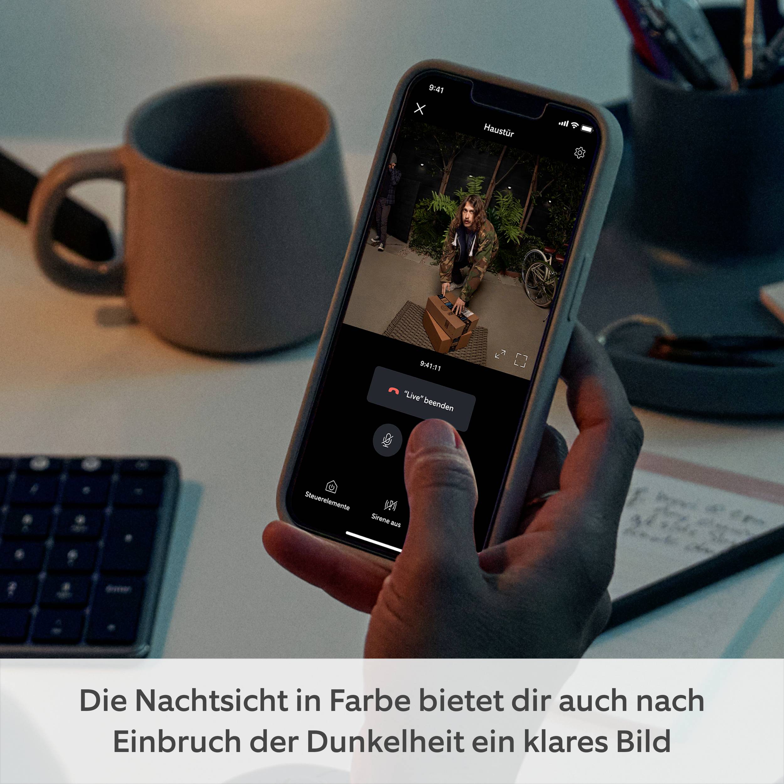 Eine Person hält ein Smartphone mit einer Live-Überwachungskameraansicht. Text am unteren Rand: 'Die Nachtsicht in Farbe bietet dir auch nach Einbruch der Dunkelheit ein klares Bild'.
