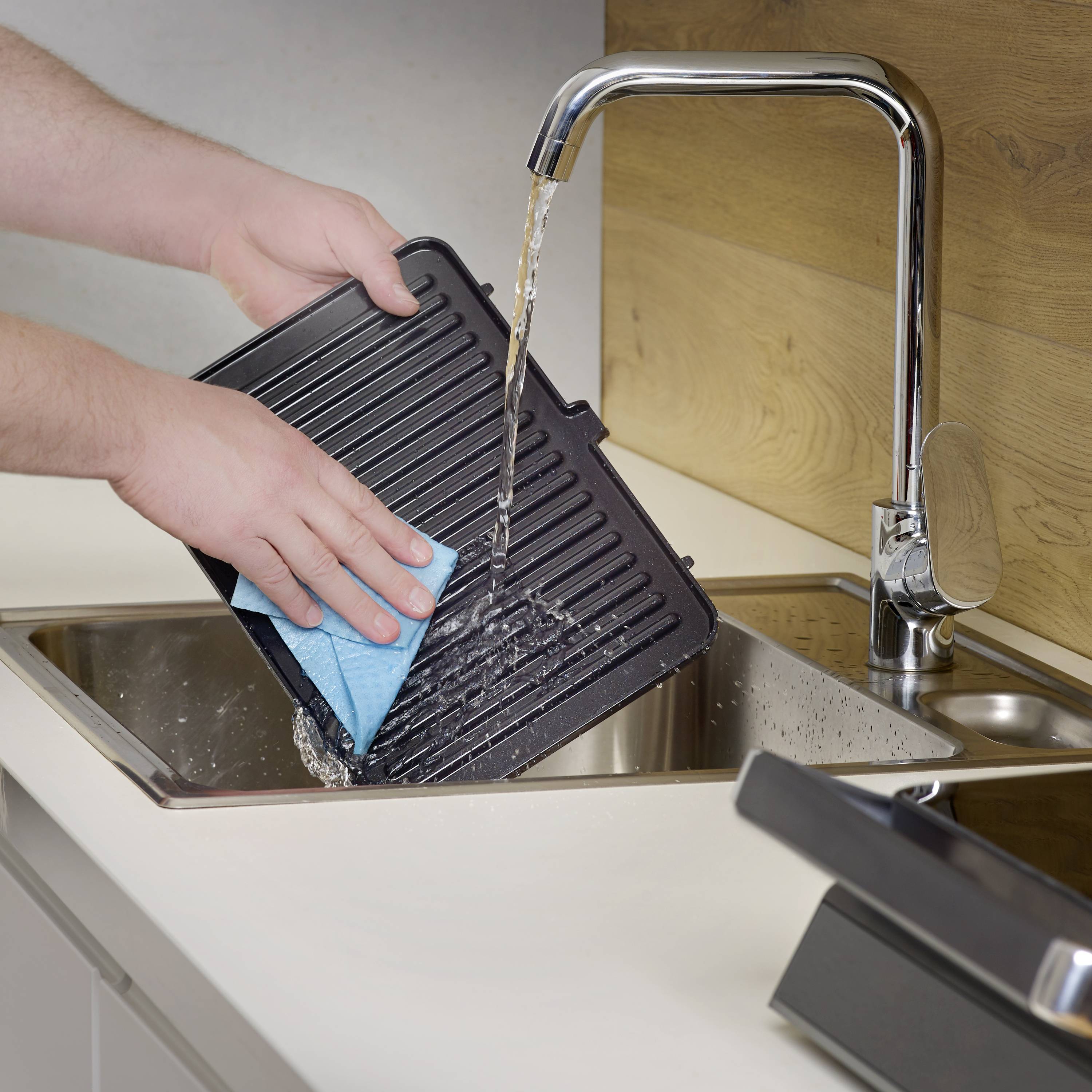 Eine Person spült ein Grillblech mit einem blauen Schwamm in einem Waschbecken, während Wasser aus dem Wasserhahn läuft.