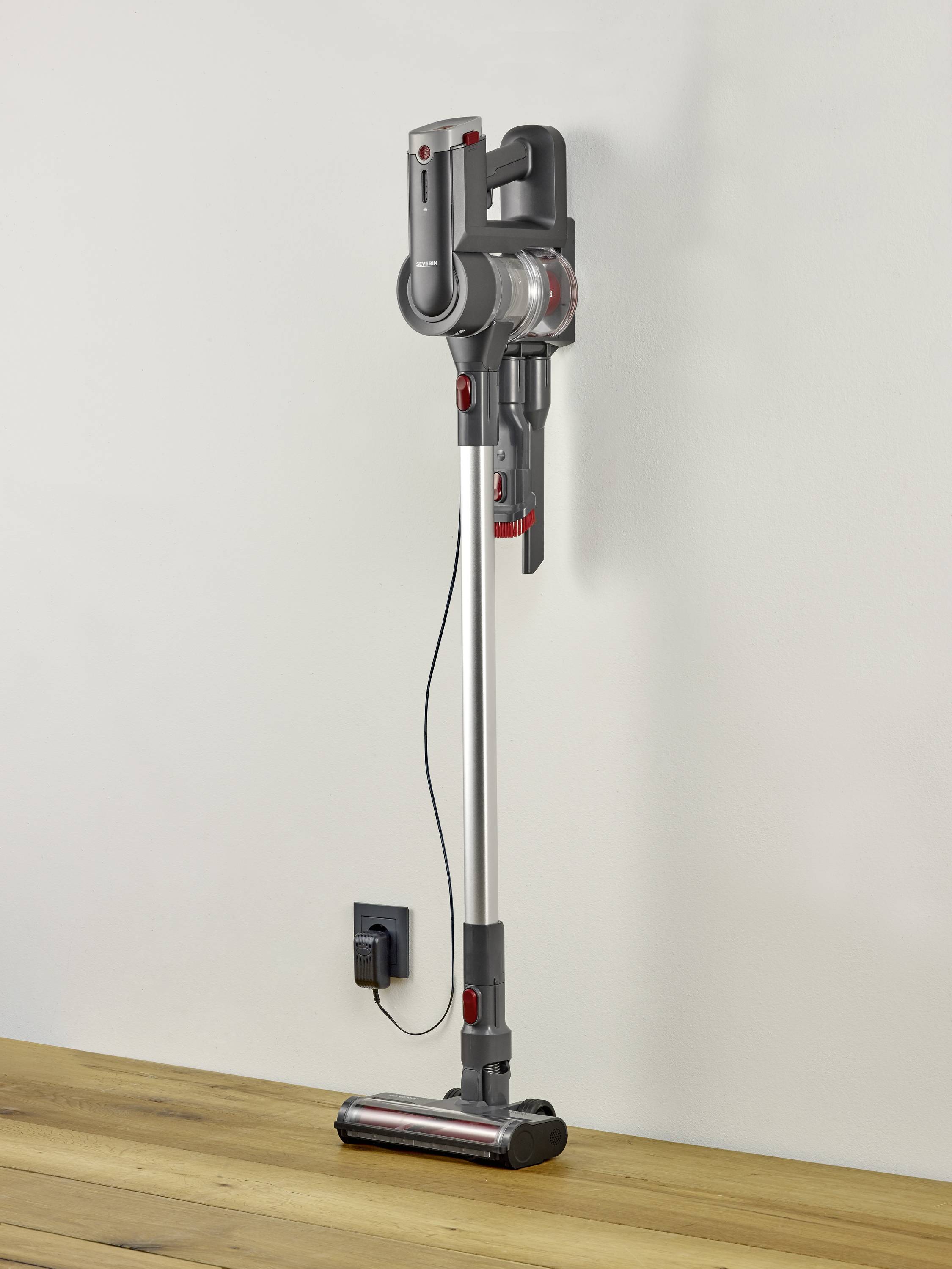 Ein kabelloser Staubsauger ist vertikal an einer weißen Wand aufgehängt. Er hat ein modernes Design mit grauen und roten Akzenten.