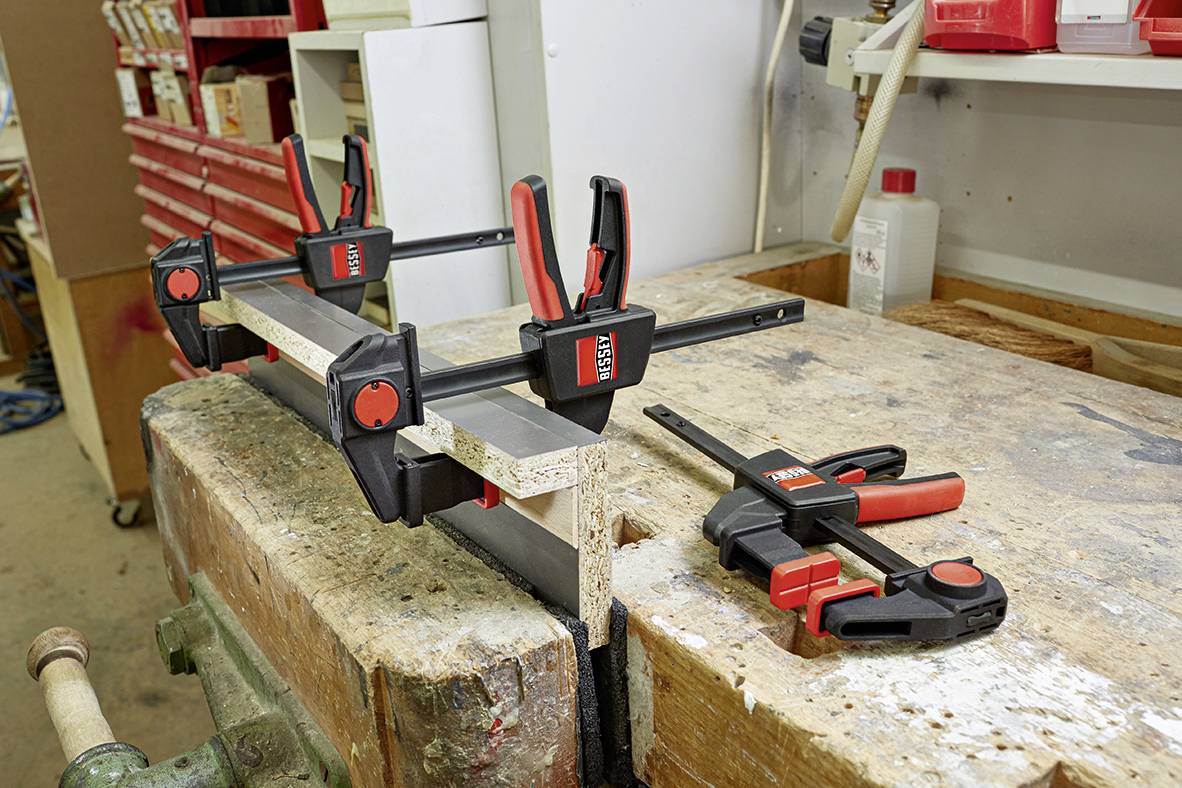 Werkbank mit Holzleiste, die mit mehreren Schraubzwingen an einem Tisch befestigt ist; eine zusätzliche Zwinge liegt daneben.