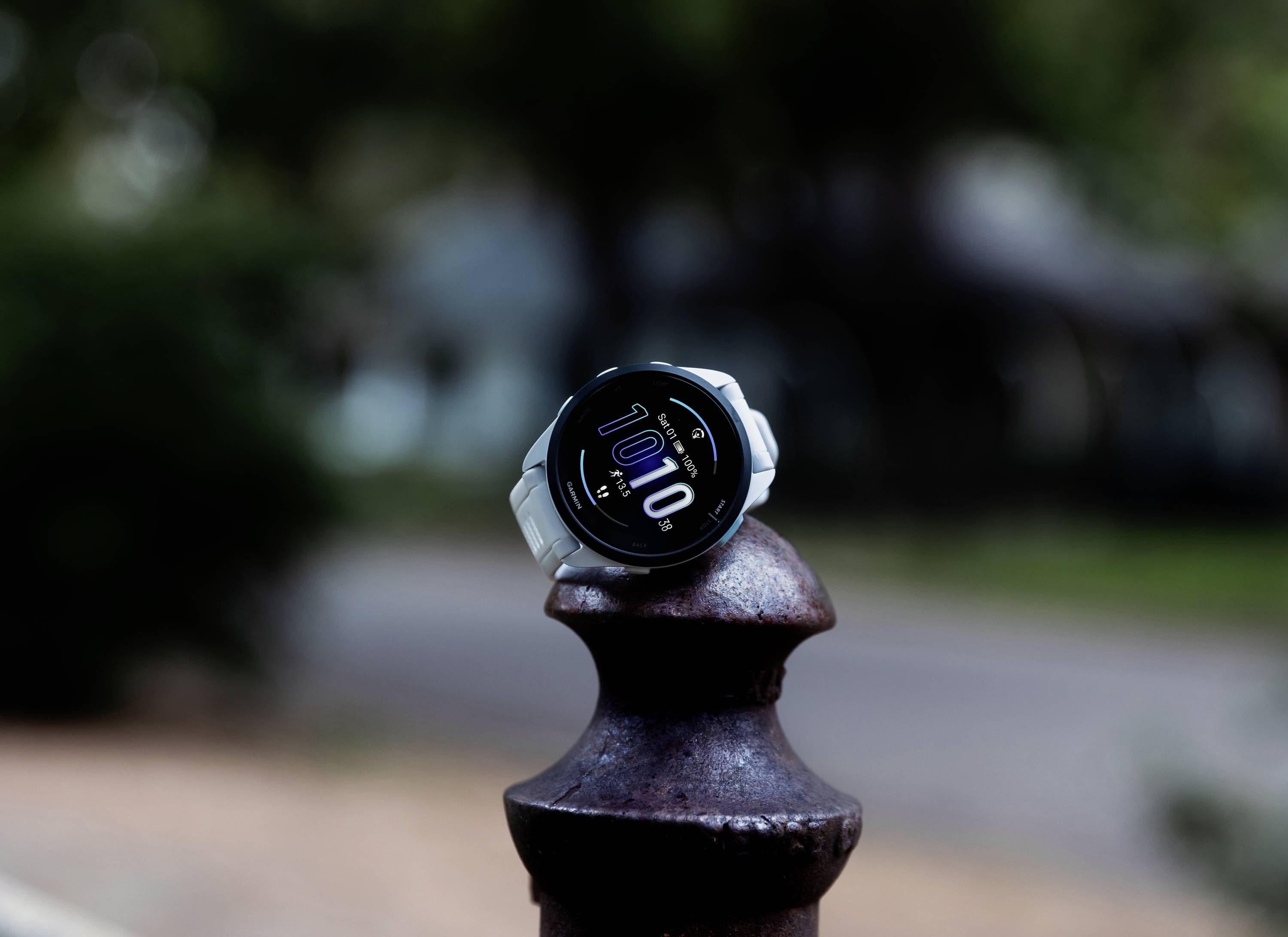 Eine Smartwatch mit weißem Armband zeigt die Uhrzeit 10:10 auf einem runden Sockel im Freien. Der Hintergrund ist verschwommen.