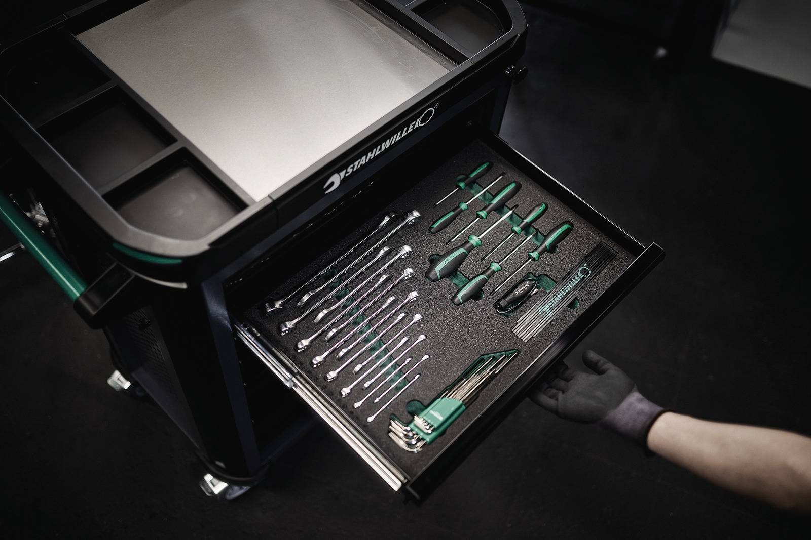 Ein geöffneter Werkstattwagen mit einer Schublade voller ordentlicher grüner Werkzeuge. Eine behandschuhte Hand zieht die Schublade heraus.