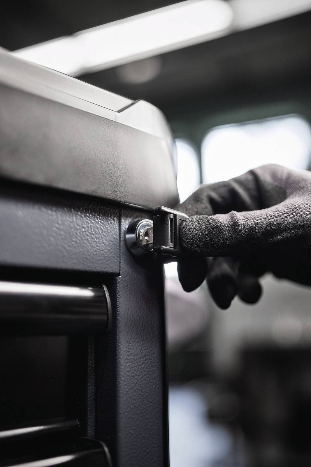 Hand mit schwarzem Handschuh verriegelt einen Schrank in einer Werkstatt. Im Hintergrund sind unscharfe Werkzeuge sichtbar.