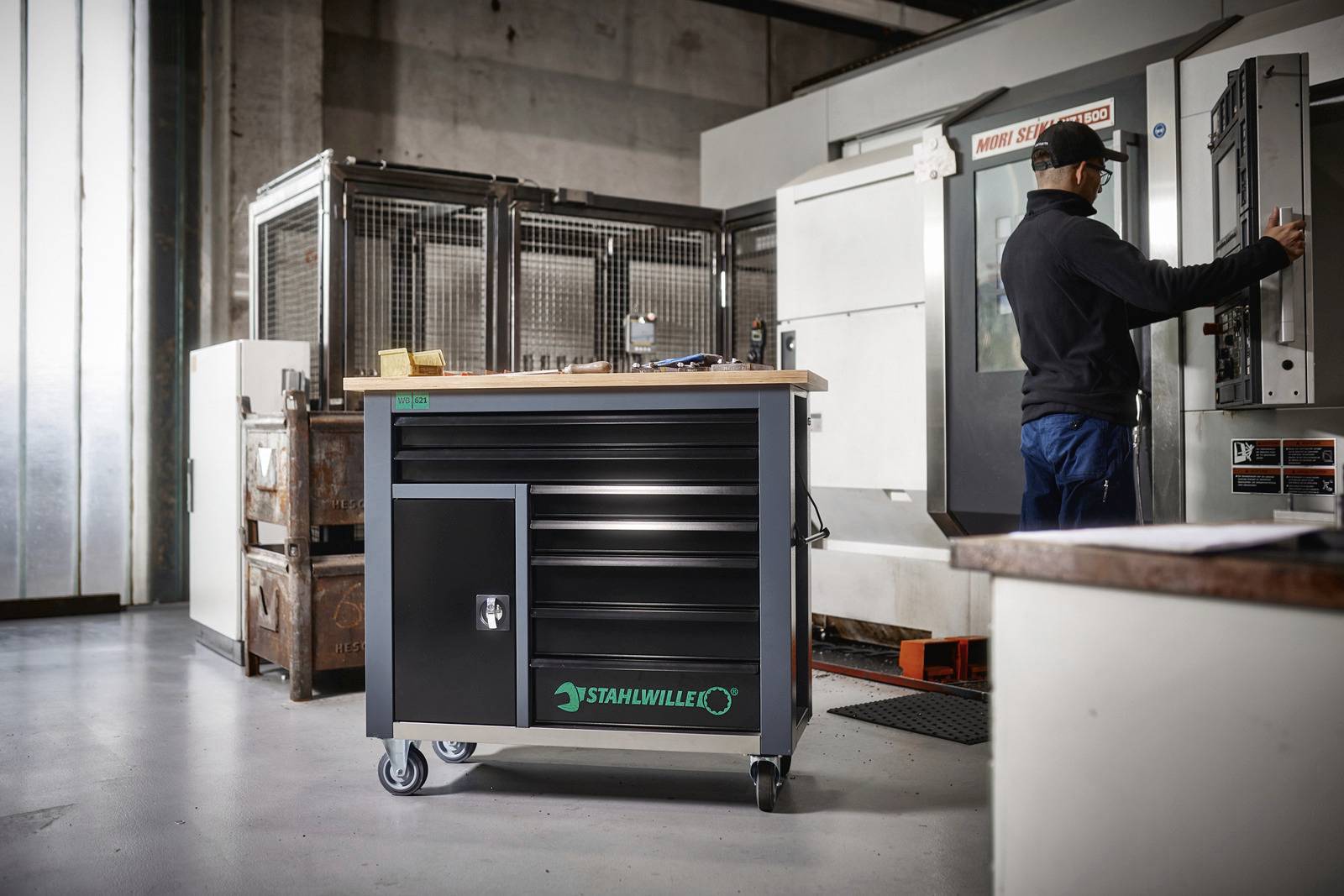 Rollwagen mit Markennamen, steht in einer Werkstatt. Im Hintergrund bedient eine Person eine Maschine. Industrielle Umgebung.