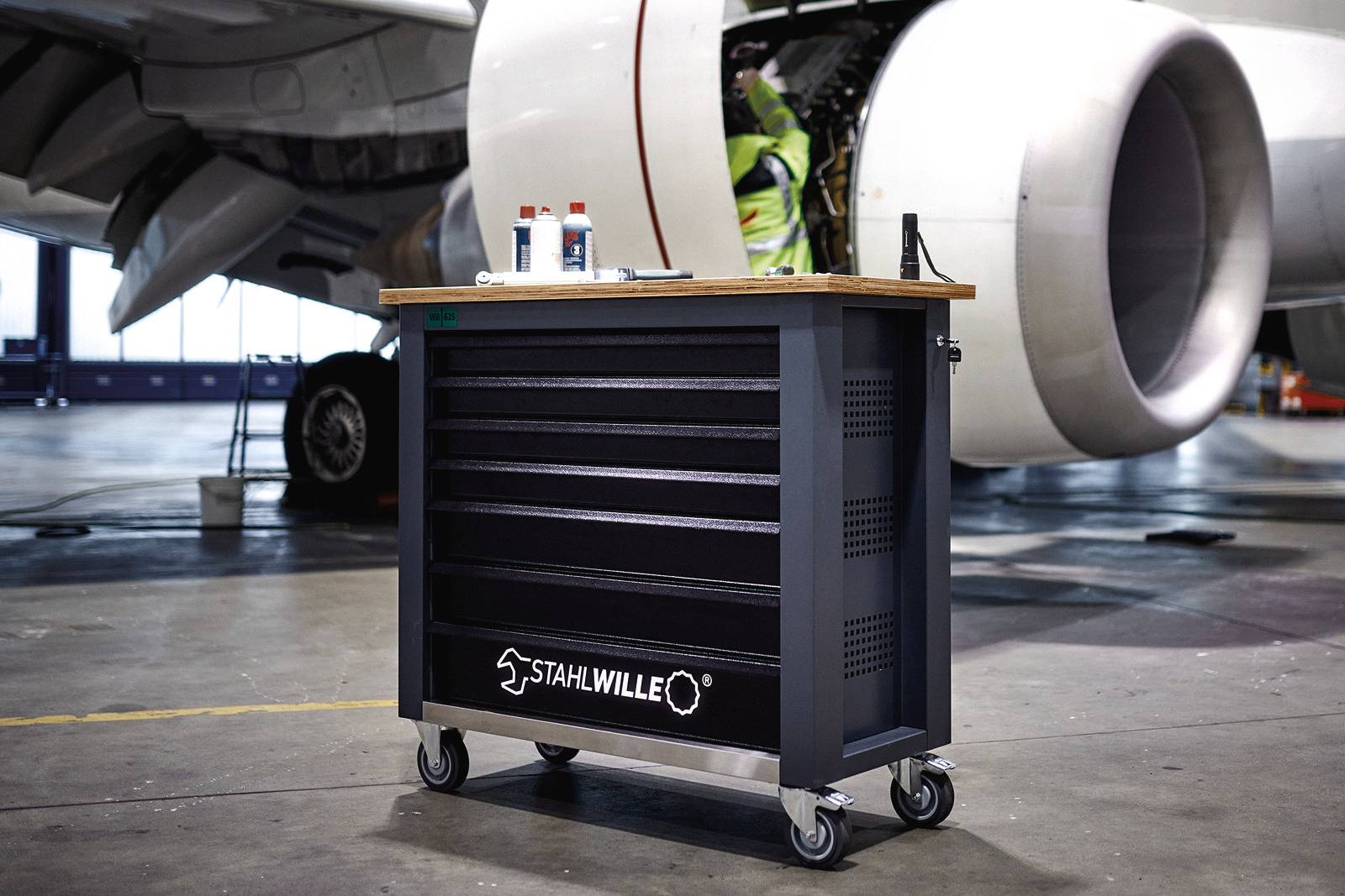 Werkzeugwagen auf Rädern steht in einer Flugzeugwerkstatt. Im Hintergrund ist ein Flugzeugtriebwerk und ein Techniker arbeitend sichtbar.