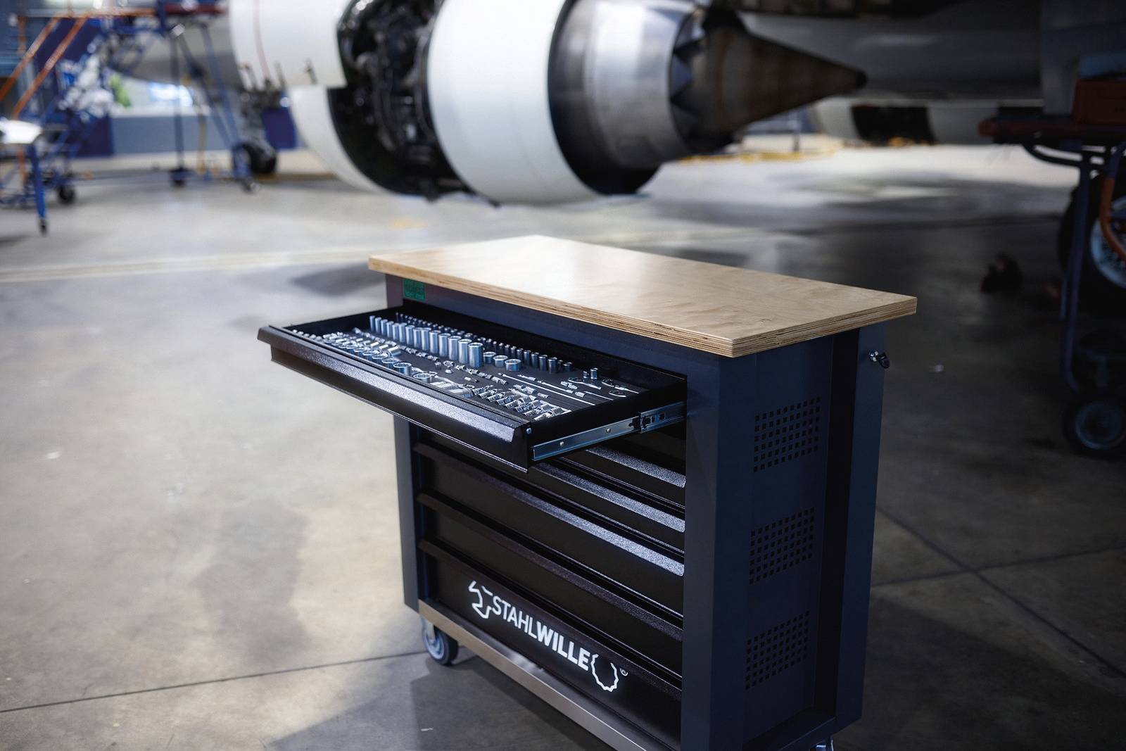 Ein Werkzeugwagen mit offenen Schubladen, gefüllt mit Werkzeugen, steht in einem Hangar vor einem Flugzeugtriebwerk.
