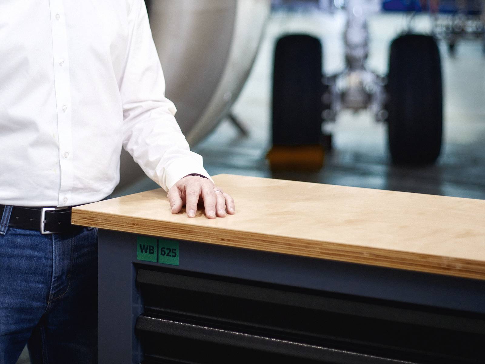 Ein Mann steht neben einer Werkbank, die in einer Werkstatt vor einem unscharfen Flugzeugfahrwerk zu sehen ist.