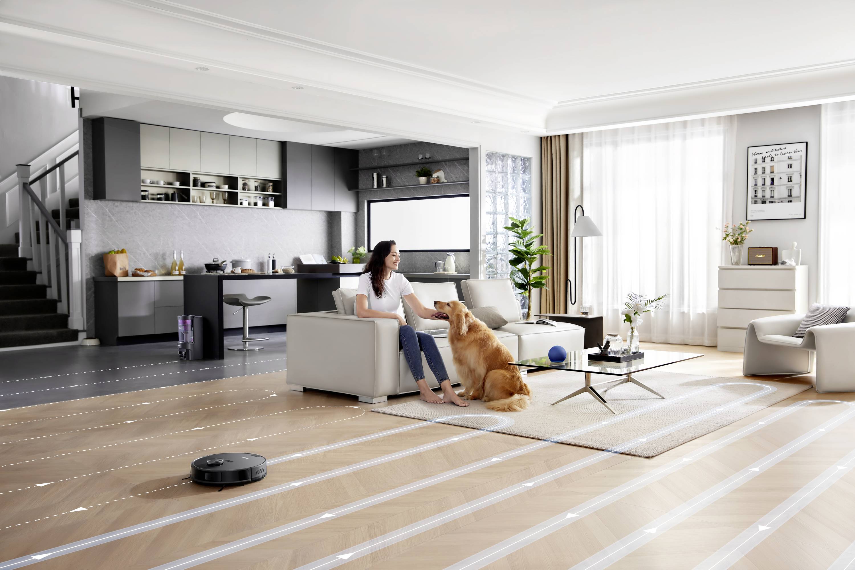 Eine Frau sitzt auf dem Sofa und streichelt einen Hund in einem modernen Wohnzimmer mit offener Küche. Ein Saugroboter reinigt den Boden.