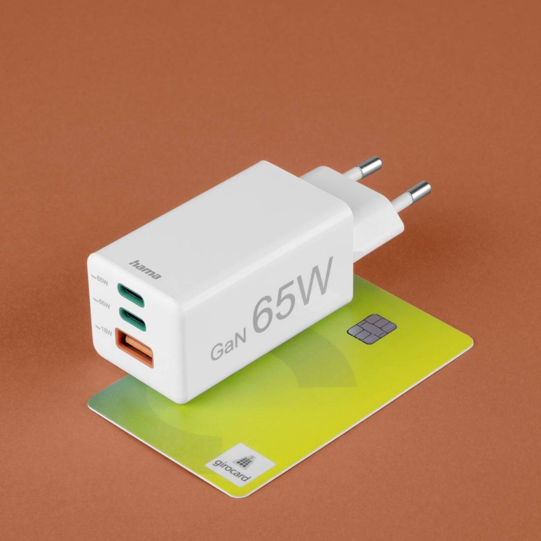 Ein weißes 65W GaN-Ladegerät von 'hama' mit zwei USB-C- und einem USB-A-Anschluss liegt auf einer grünen Karte auf orangefarbenem Hintergrund.