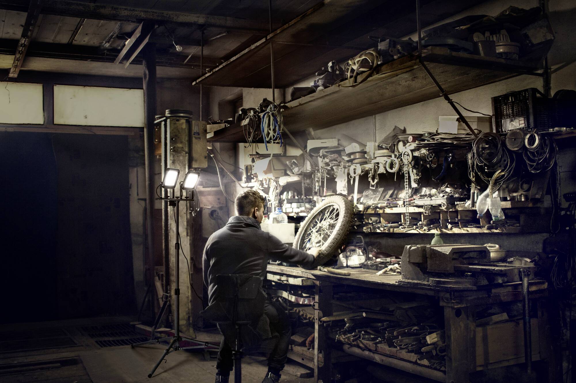 Ein Mann arbeitet in einer dunklen Werkstatt an einem Fahrrad. Hintergrund voller Werkzeuge und Ersatzteile, Beleuchtung von rechts.
