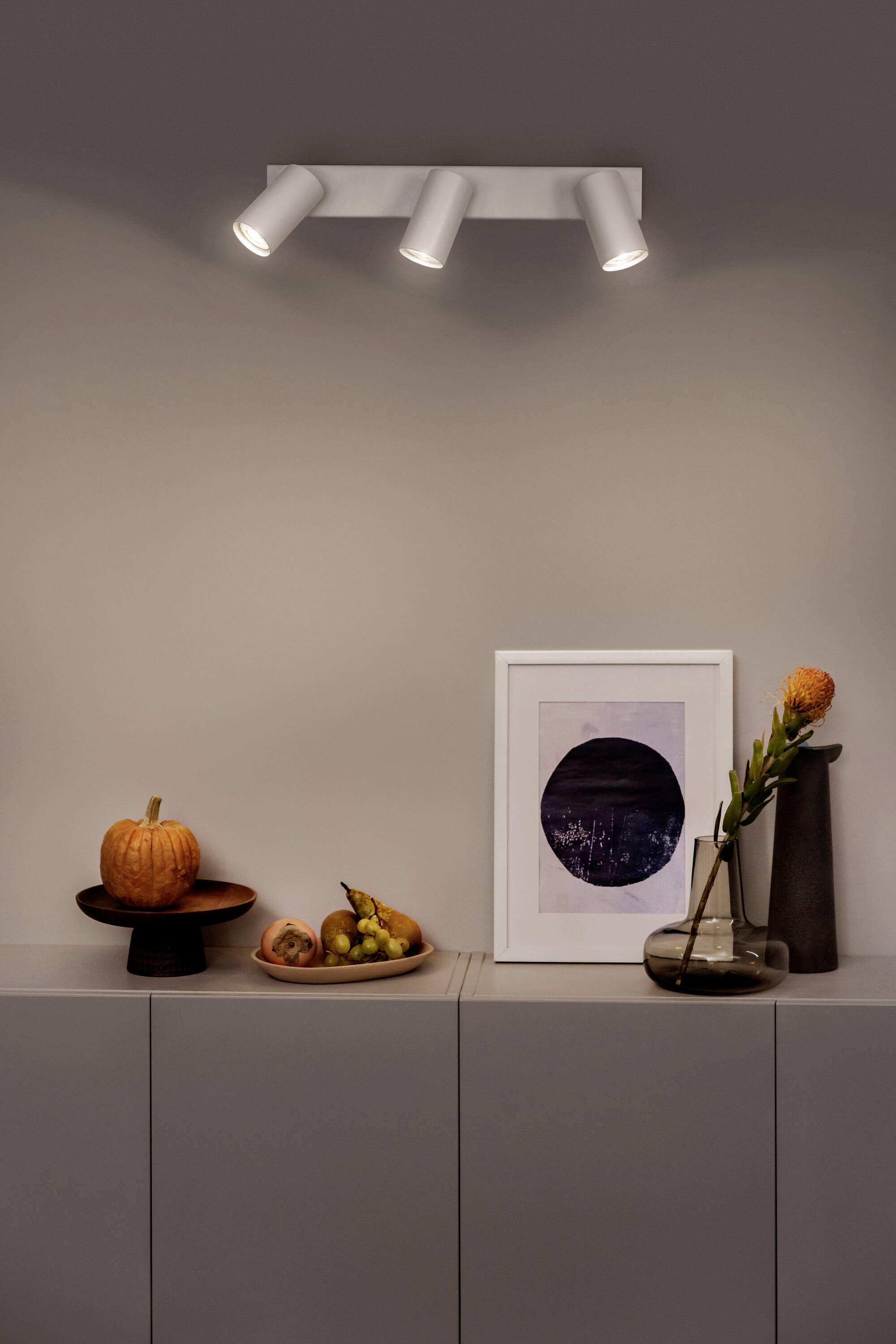Ein minimalistisches Sideboard mit Kürbissen, einem Obstteller, einer Vase mit Blume und einem gerahmten Bild darunter anmutige Beleuchtung.