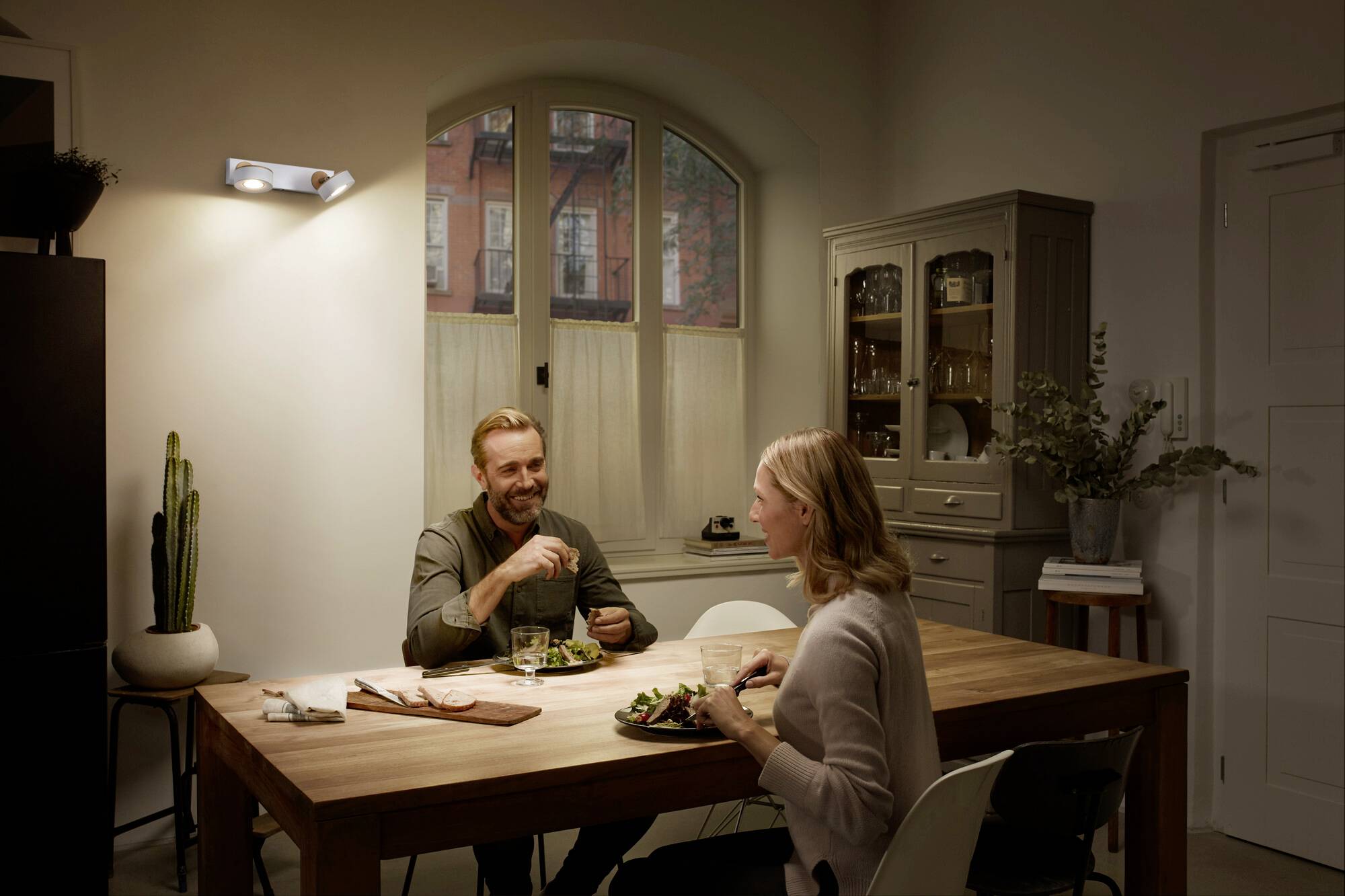 Eine Frau und ein Mann sitzen lächelnd an einem Holztisch in einer gemütlich beleuchteten Küche und essen gemeinsam.