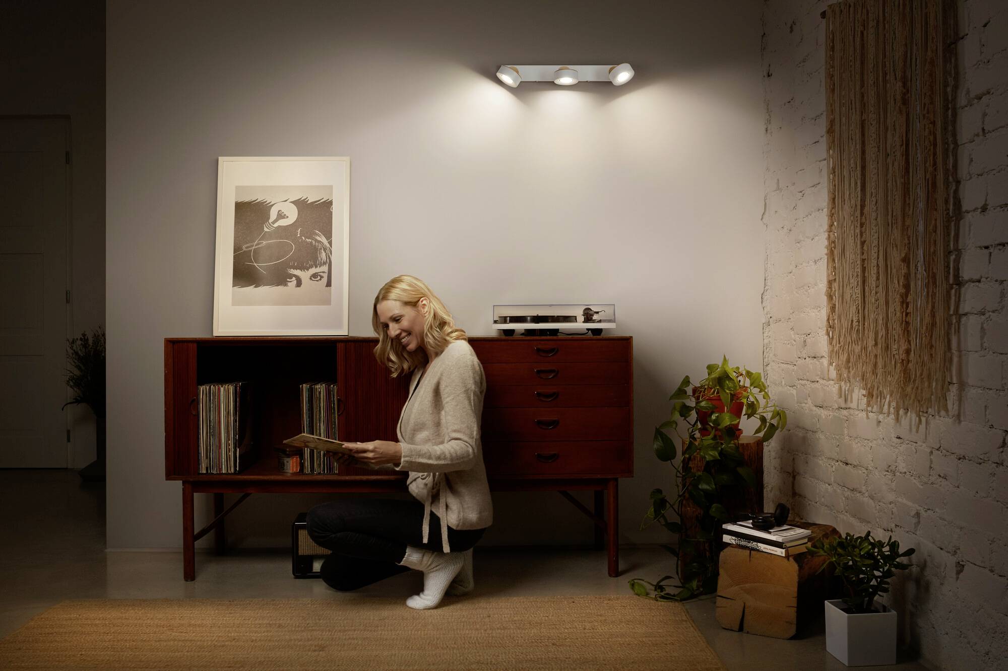 Eine Frau kniet vor einem Schrank in einem Wohnzimmer, sortiert Schallplatten. Im Hintergrund sind Pflanzen und dezente Beleuchtung.