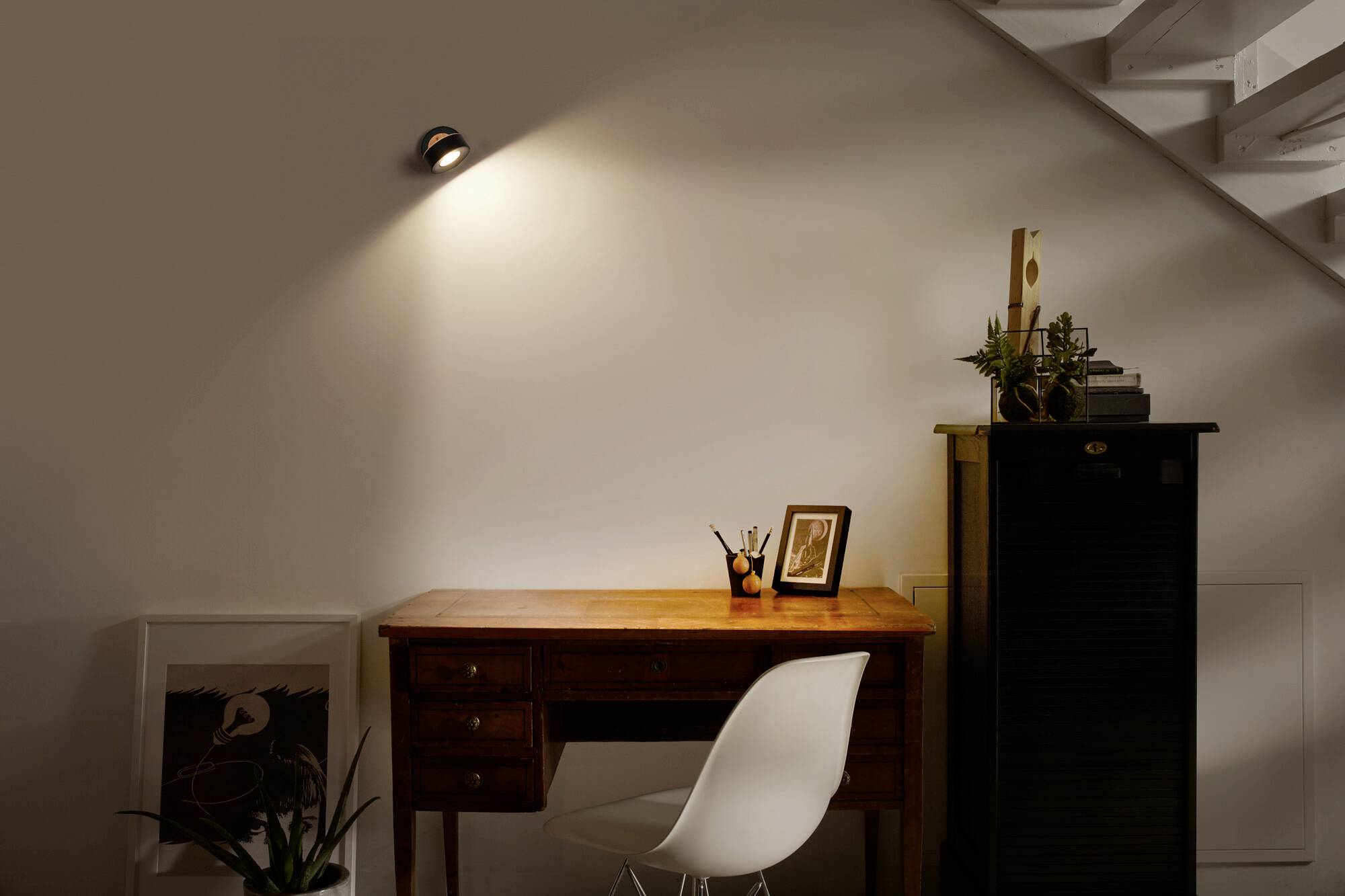 Ein Holzschreibtisch mit einem Stuhl steht unter einer Treppe. Auf dem Tisch steht ein gerahmtes Foto, einige Stifte und Pflanzen. Ein Spotlicht beleuchtet den Tisch.