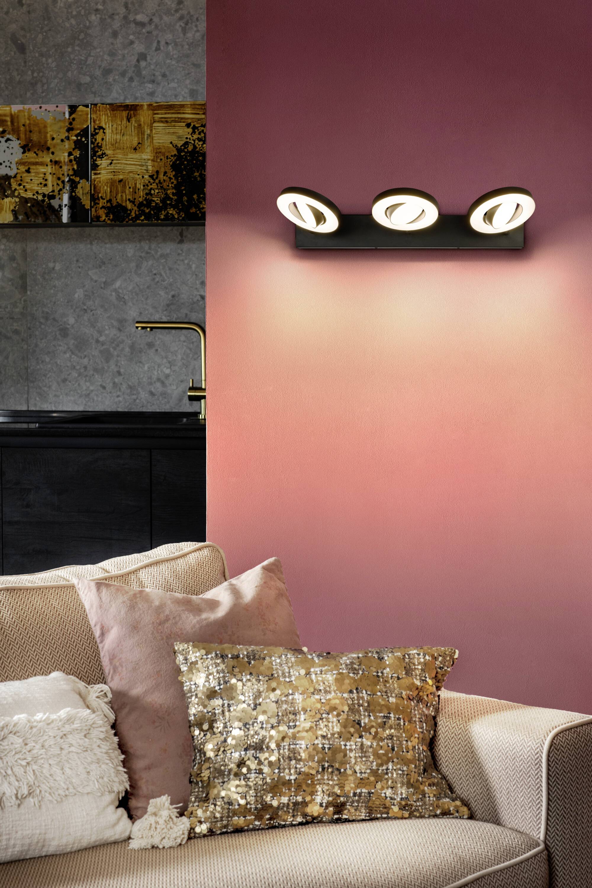 Wohnzimmer mit beigem Sofa, buntem Kissen und Wandlampe über Sofarücken. Dunkles Regal mit goldenen Dekorationen im Hintergrund.