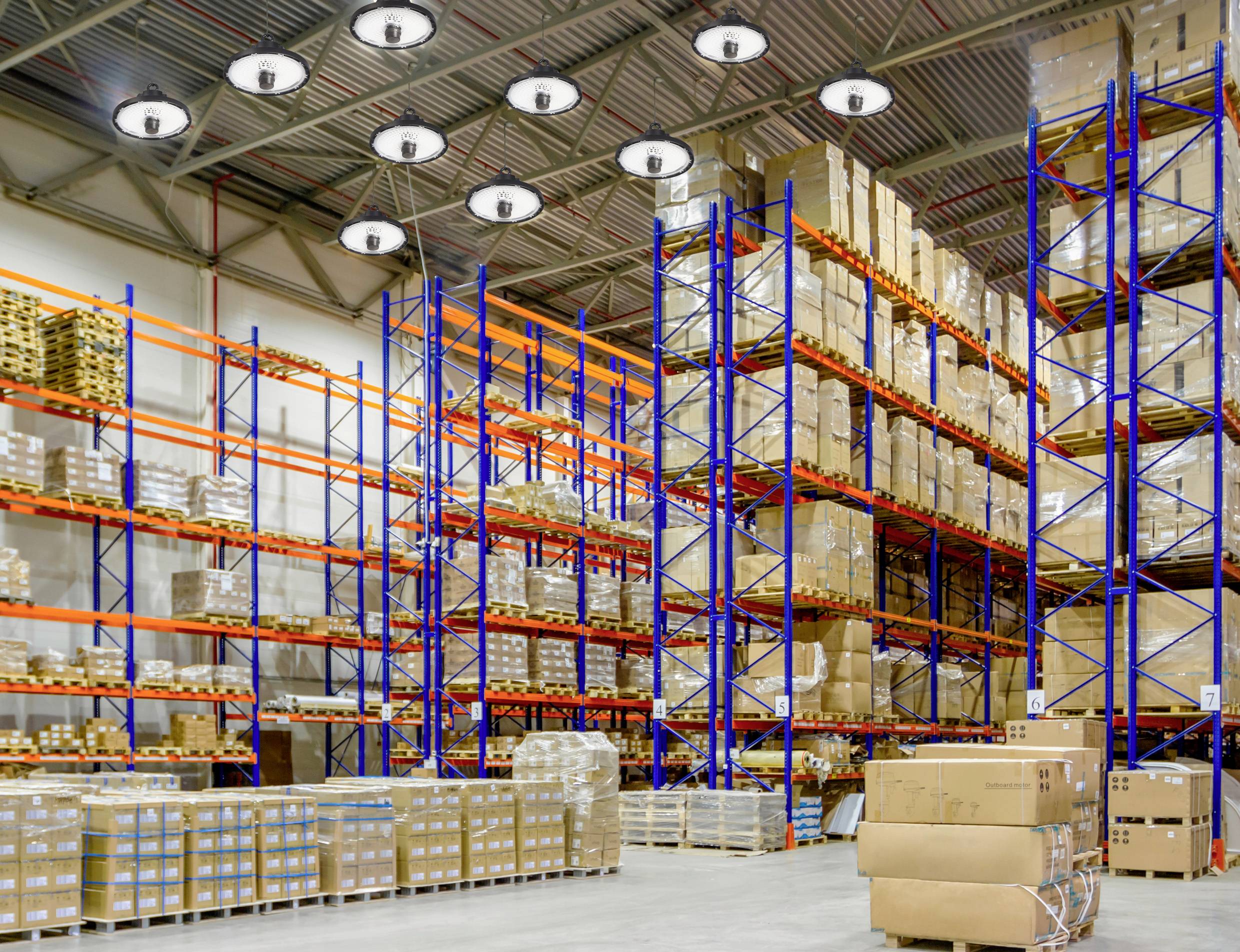 Ein großes Lager mit hohen Regalen, gefüllt mit Kisten und Paketen. Helle industrielle Beleuchtung und Paletten am Boden.