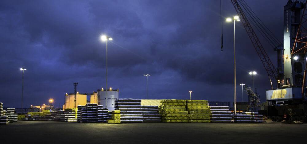 Industriegelände bei Nacht mit gestapelten Metallstangen und Baummaterial auf einem beleuchteten Hafen. Ein Kran ist im Hintergrund sichtbar.