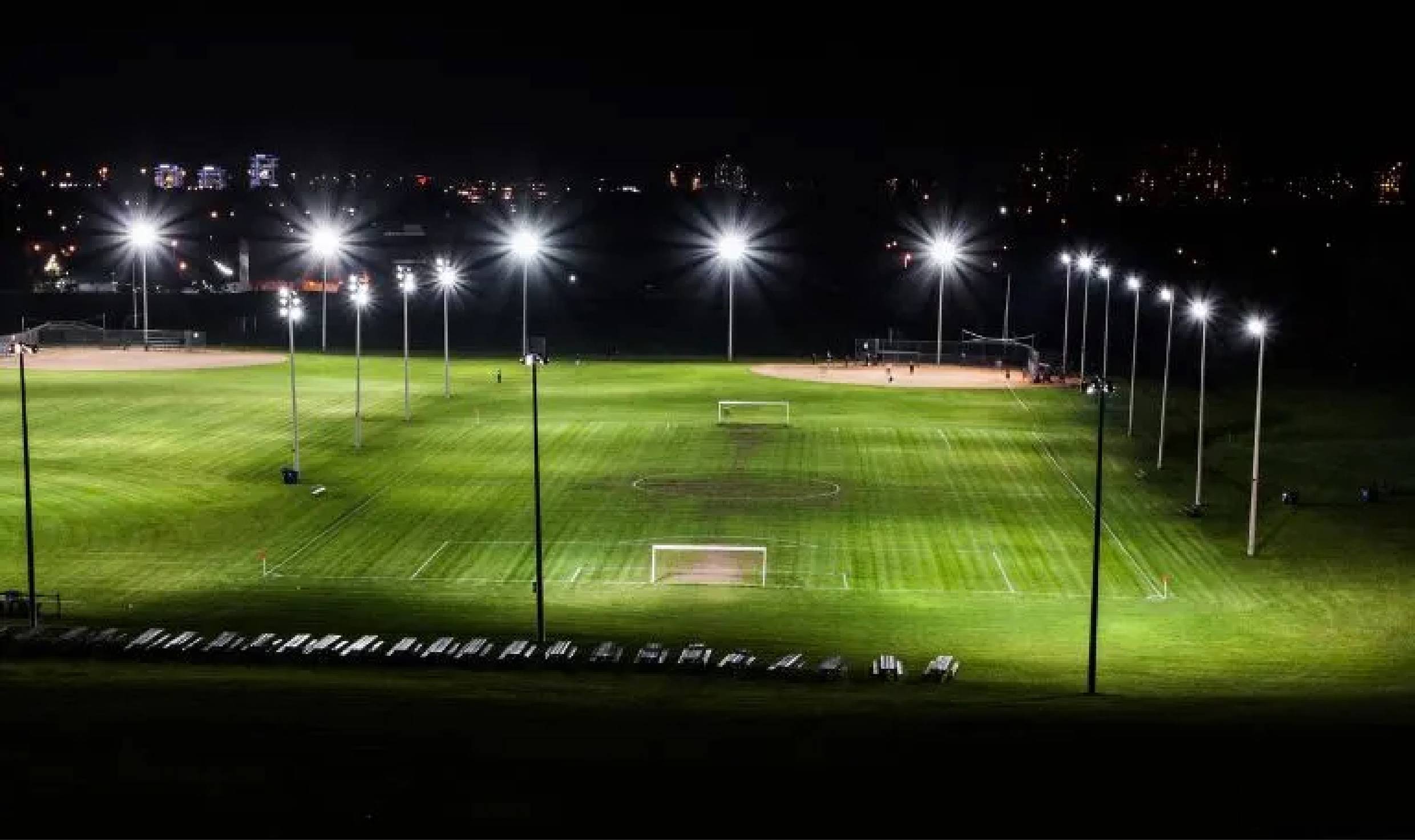 Nachts beleuchtetes Fußballfeld mit Toren, umgeben von Flutlichtmasten. Im Hintergrund sind Stadtlichter sichtbar.