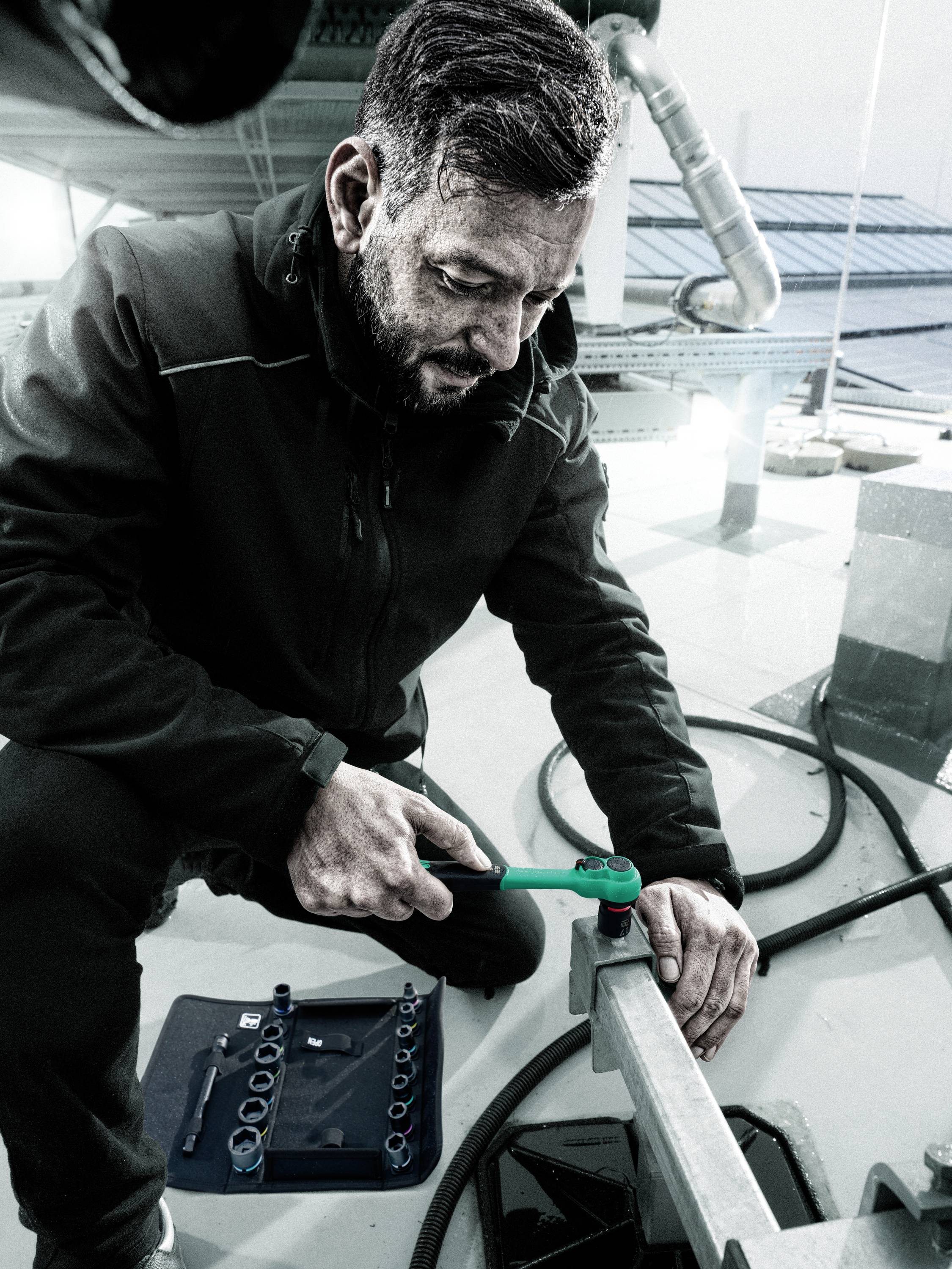 Ein Mann in Arbeitskleidung benutzt einen Schraubenschlüssel auf einem Dach. Neben ihm liegt eine Werkzeugkiste. Hintergrund: Industriegelände.