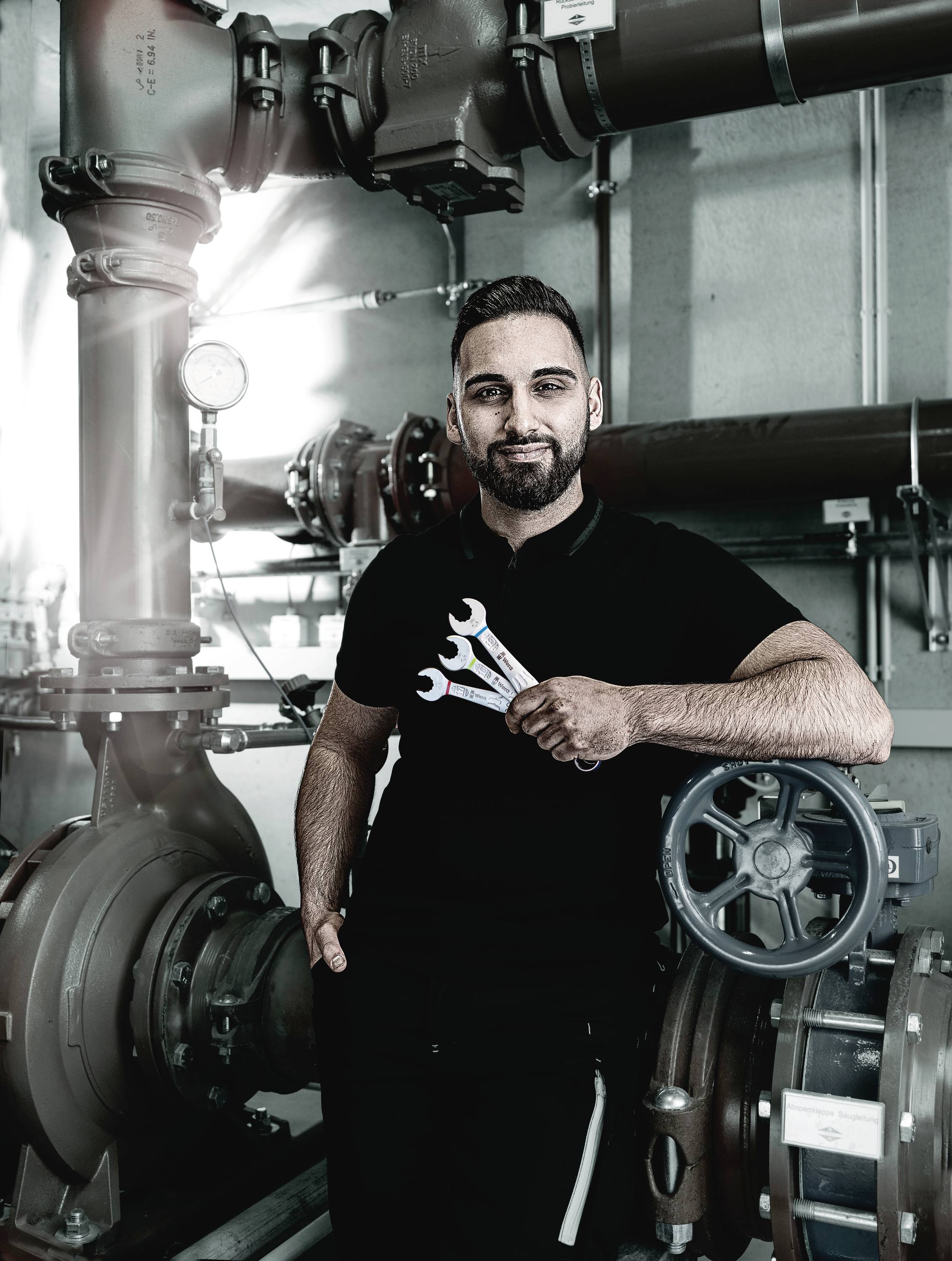 Ein Mann steht in einer industriellen Umgebung und hält drei Schraubenschlüssel. Im Hintergrund sind Rohrleitungen und Ventile zu sehen.