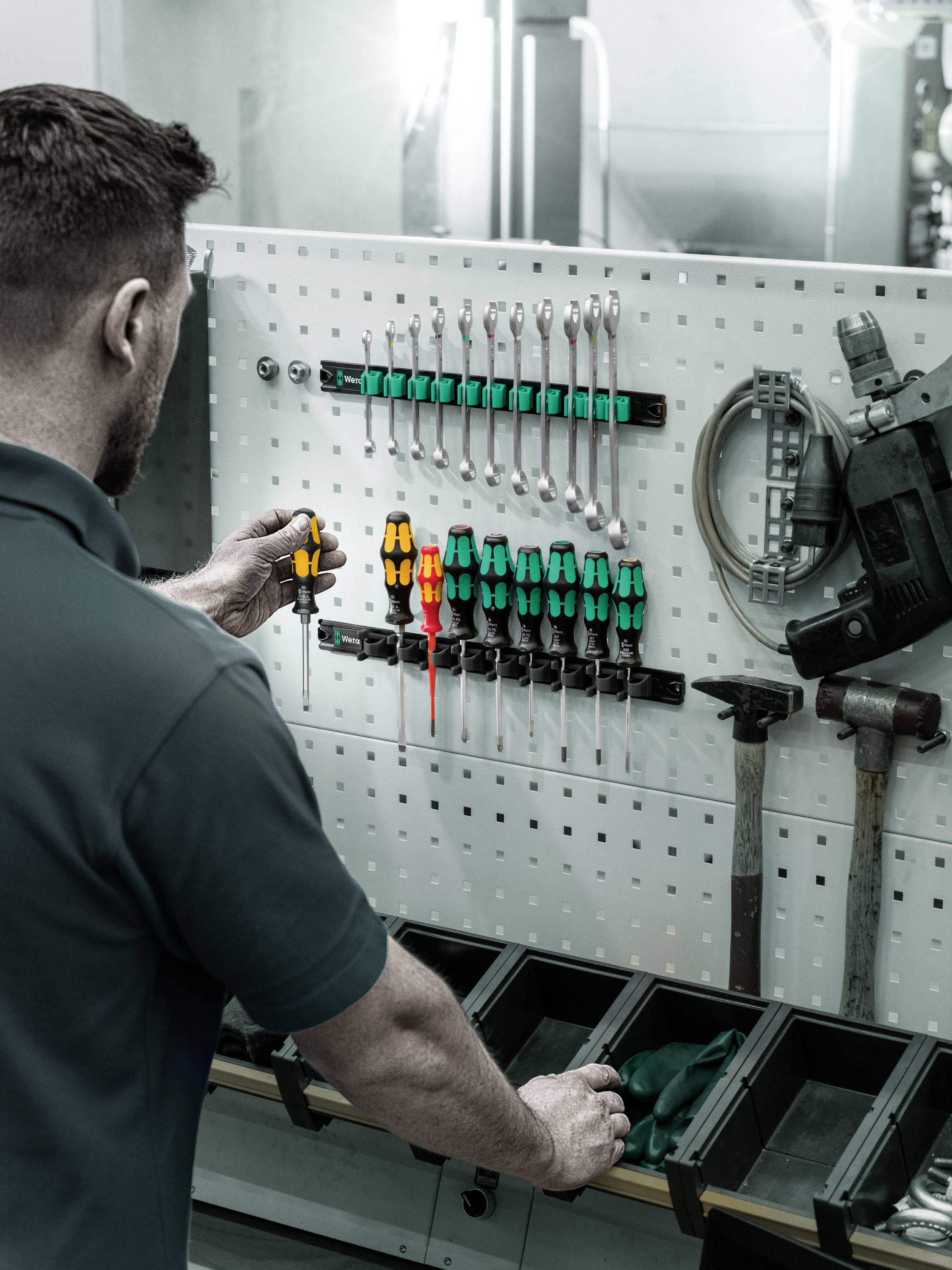 Ein Mann ordnet Werkzeuge an einer Lochwand, darunter Schraubendreher, Schraubenschlüssel und ein Elektrowerkzeug, in einer Werkstatt.