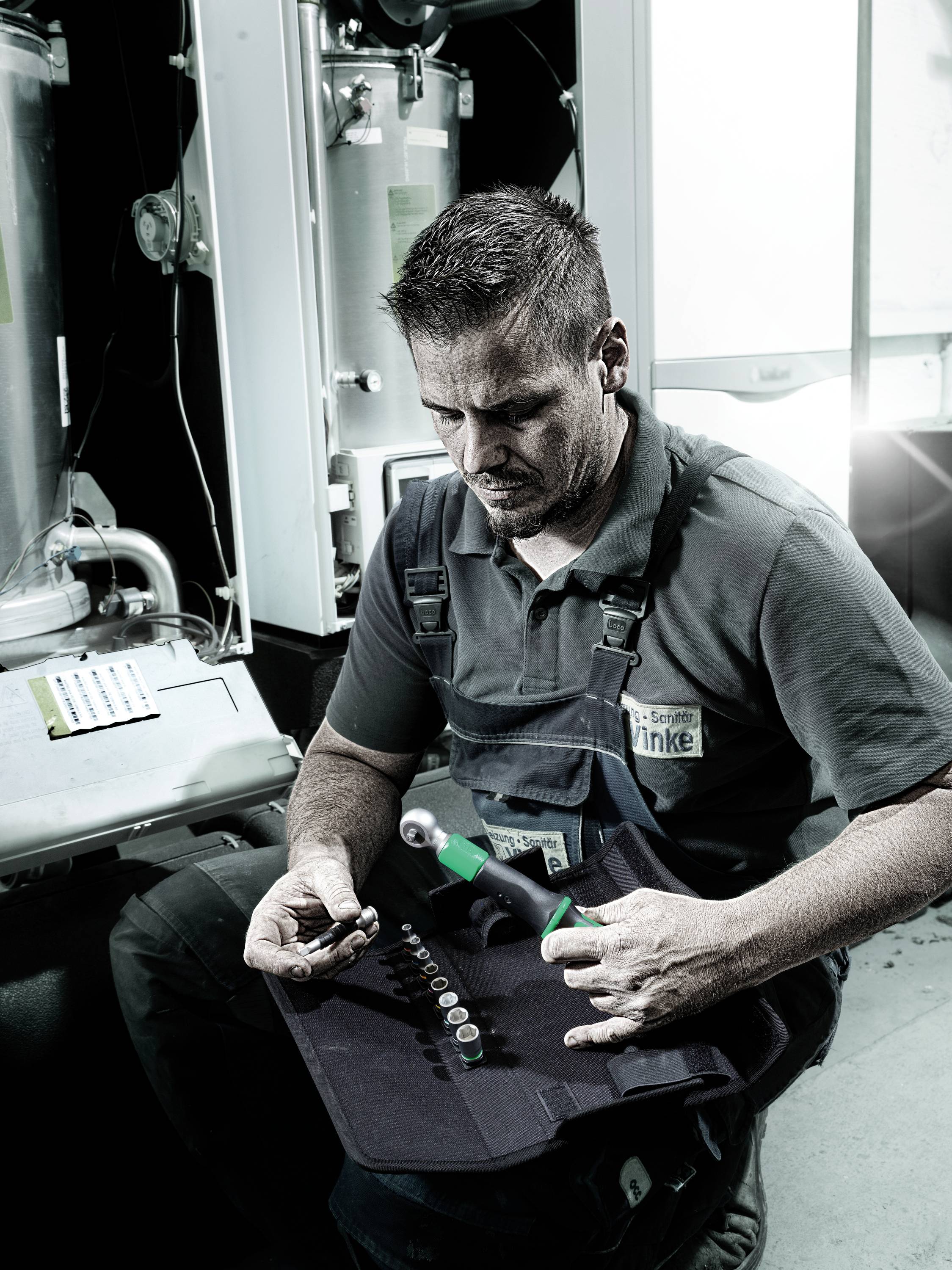 Ein Handwerker sitzt in einem Arbeitsbereich und hält Werkzeug. Im Hintergrund sind industrielle Maschinen sichtbar.